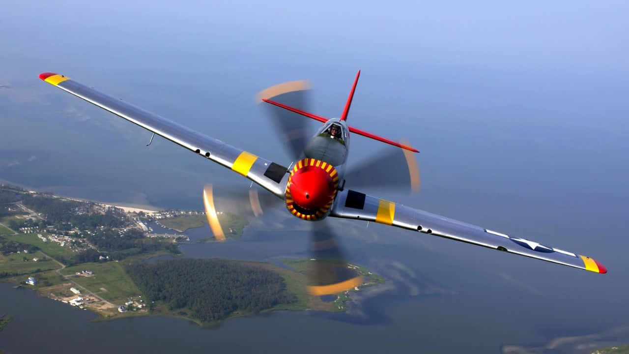 720p Plane 桌布 壁紙 — 一架紅色機頭、黃色翼尖的復古軍用戰鬥機在沿海水域和綠色島嶼上空低飛。