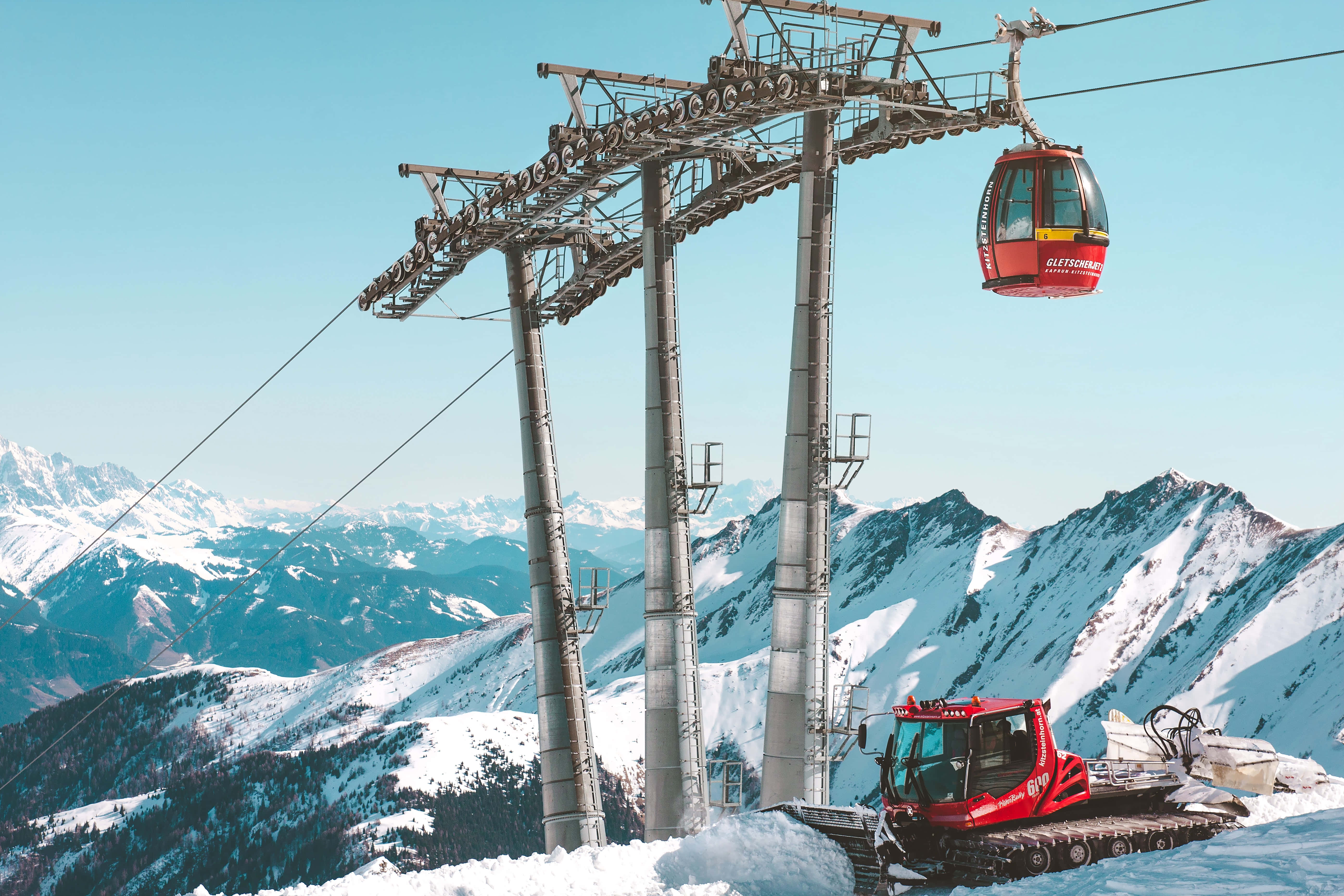 Ski Mountain 桌布 壁紙 — 一架紅色纜車在白雪覆蓋的山峰上方滑行，下方一輛紅色雪地車在晴朗的藍天下休息。