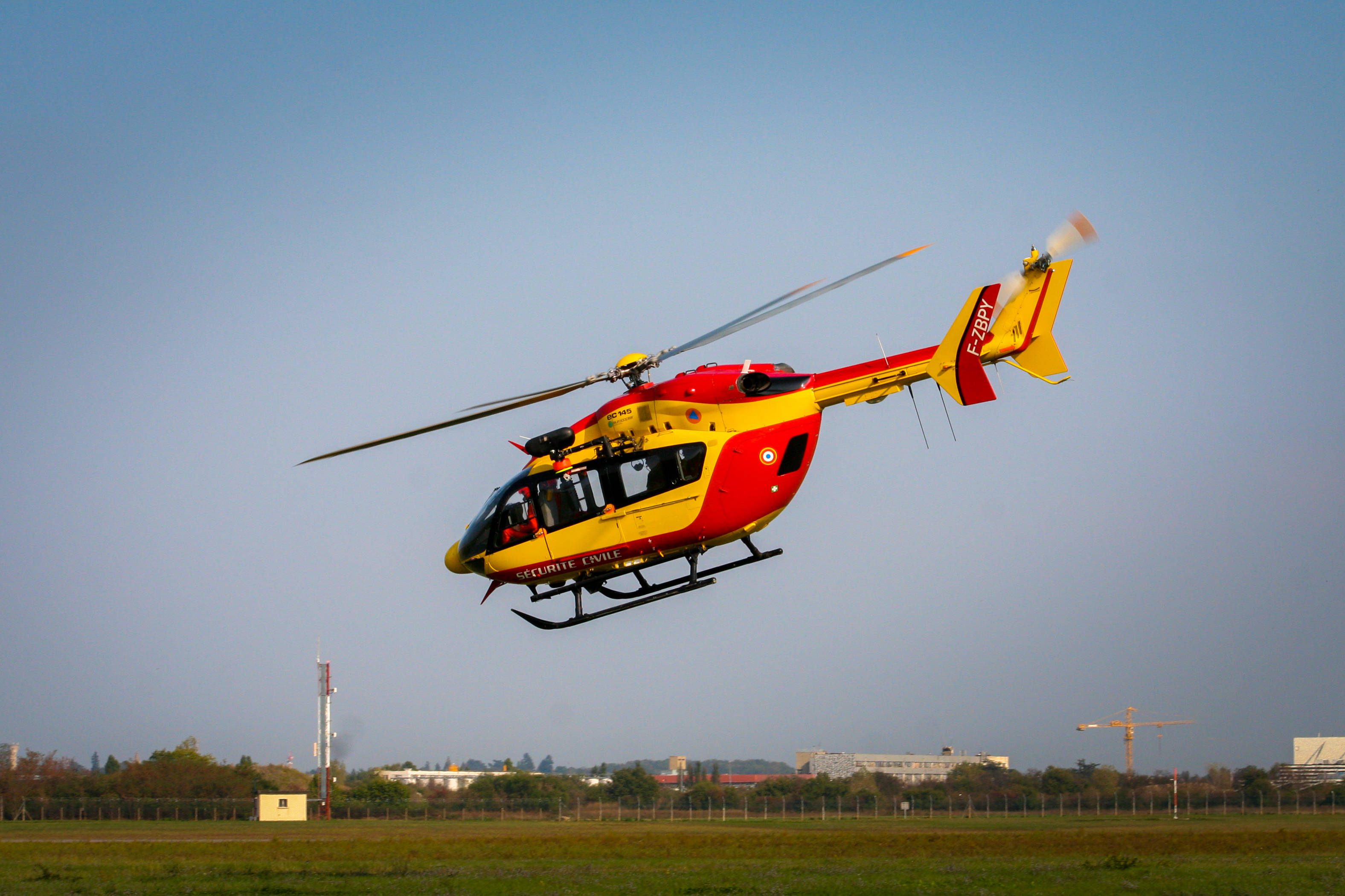 helicopter 4K 桌布 壁紙 — 一架紅黃色救援直升機在晴朗藍天下，從一片草地起飛，遠處有城市建築。