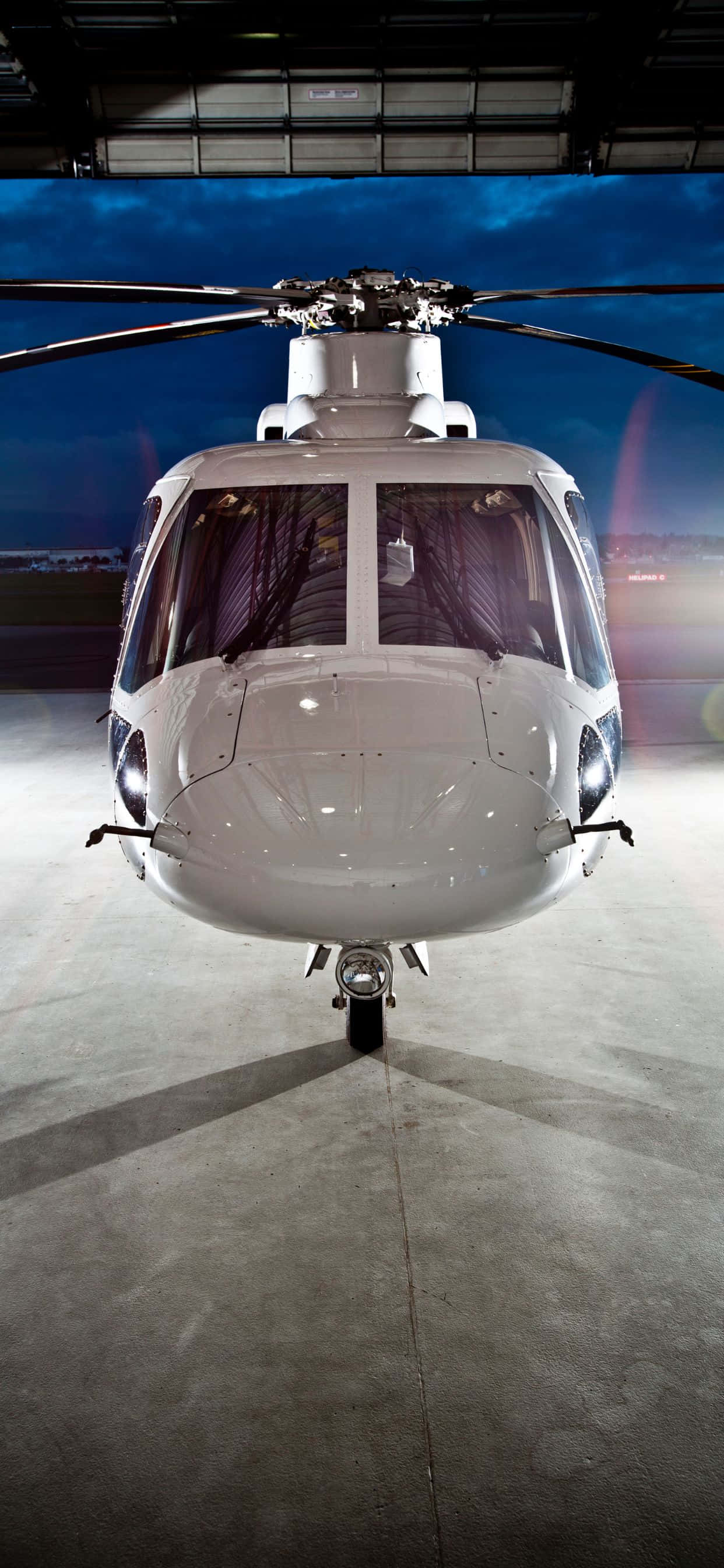 iPhone Xs Max Helicopters 桌布 壁紙 — 一架純白色直升機在機庫內向前，帶有戲劇性的藍色暮光照明。