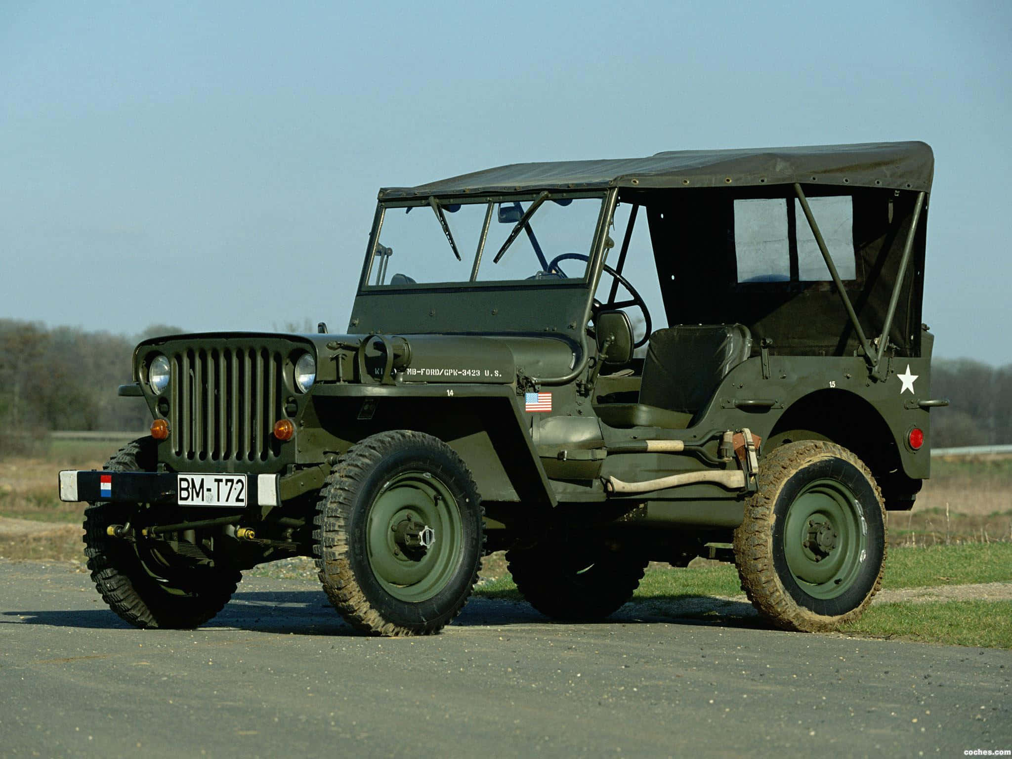 Jeep Willys 桌布 壁紙 — 一架經典橄欖綠色軍用吉普車停泊在鋪砌道路上，周圍有草地和樹木，天空清澈湛藍。