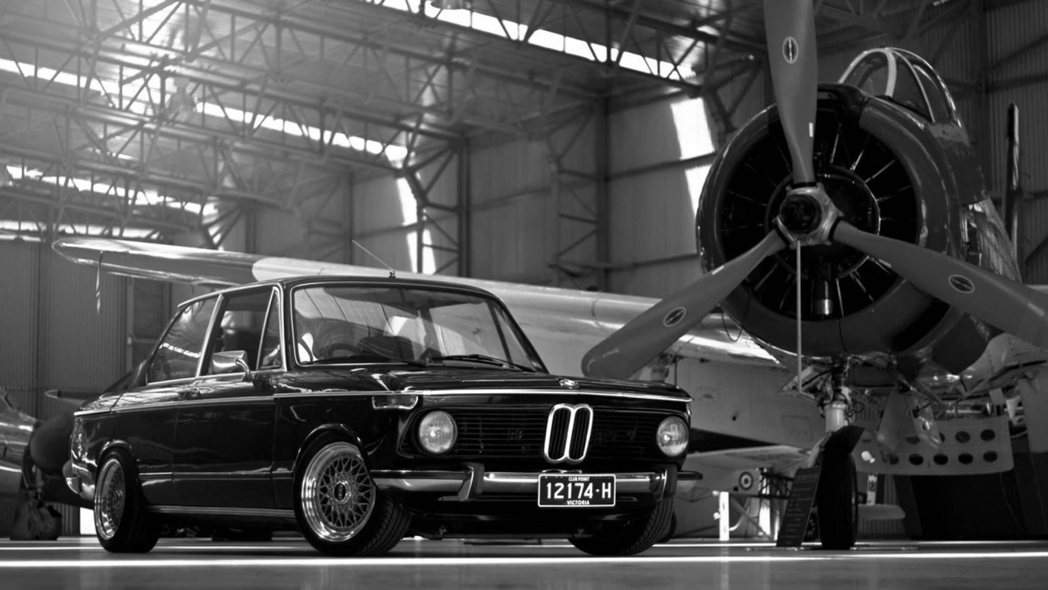 classic bmw 桌布 壁紙 — 一架經典黑色寶馬轎車停放在寬敞機庫內，旁邊是一架復古螺旋槳飛機。