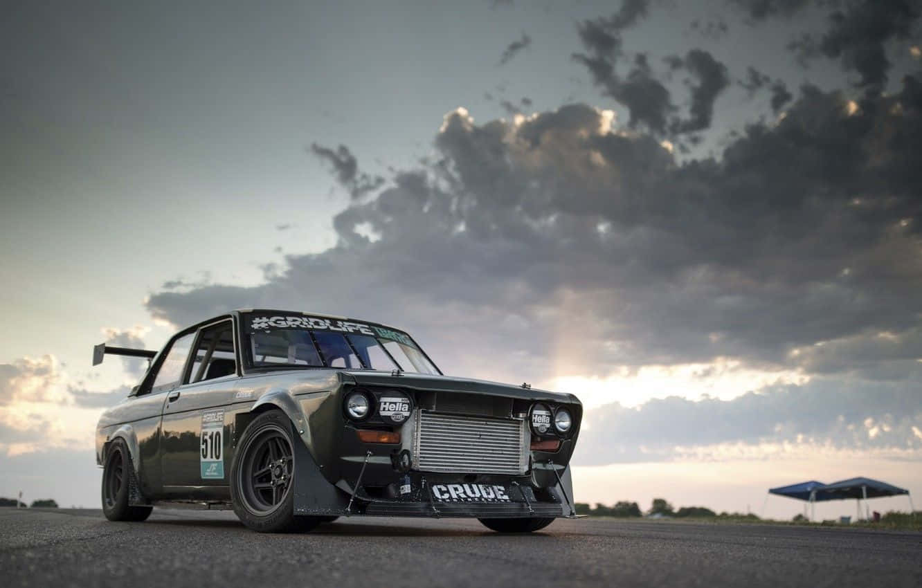 Datsun 桌布 壁紙 — 一架經過改裝的深綠色賽車，帶有 'CRUDE' 品牌標誌，在黃昏時分戲劇性的多雲天空下，停在瀝青賽道上。