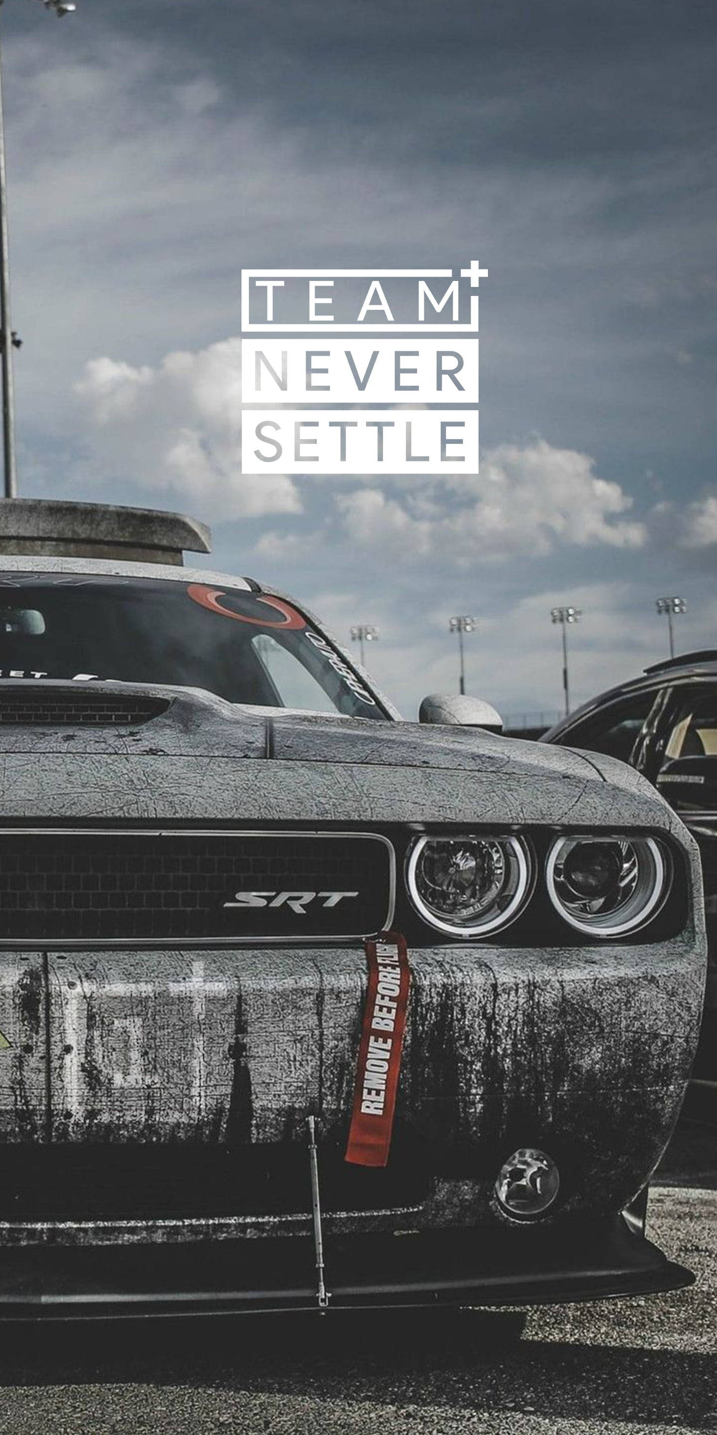 語錄 桌布 壁紙 — 一架經過風化處理的黑色道奇挑戰者SRT，帶有「TEAM NEVER SETTLE」字樣，停泊在陰天下的賽車場上。