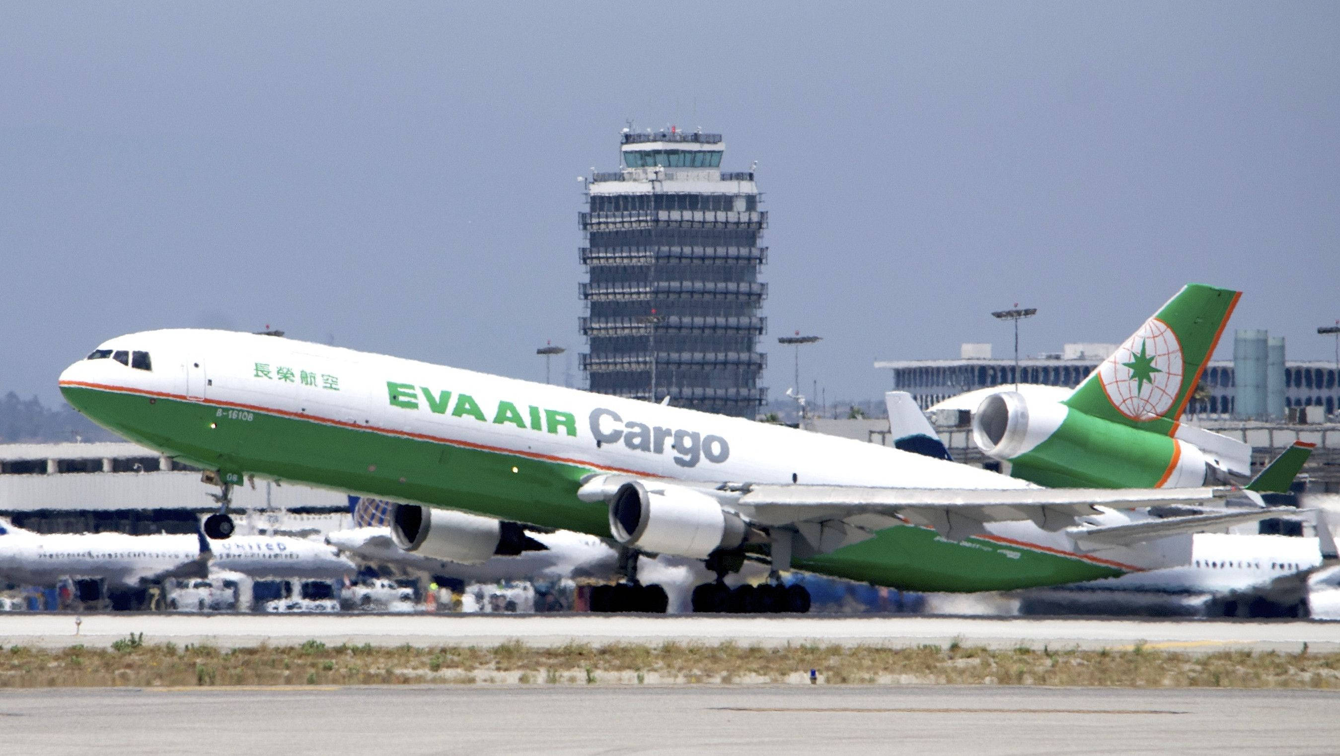 桌布 壁紙 — 一架綠色和白色的EVA Air Cargo飛機在陽光明媚的機場停機坪上滑行，背景是控制塔。