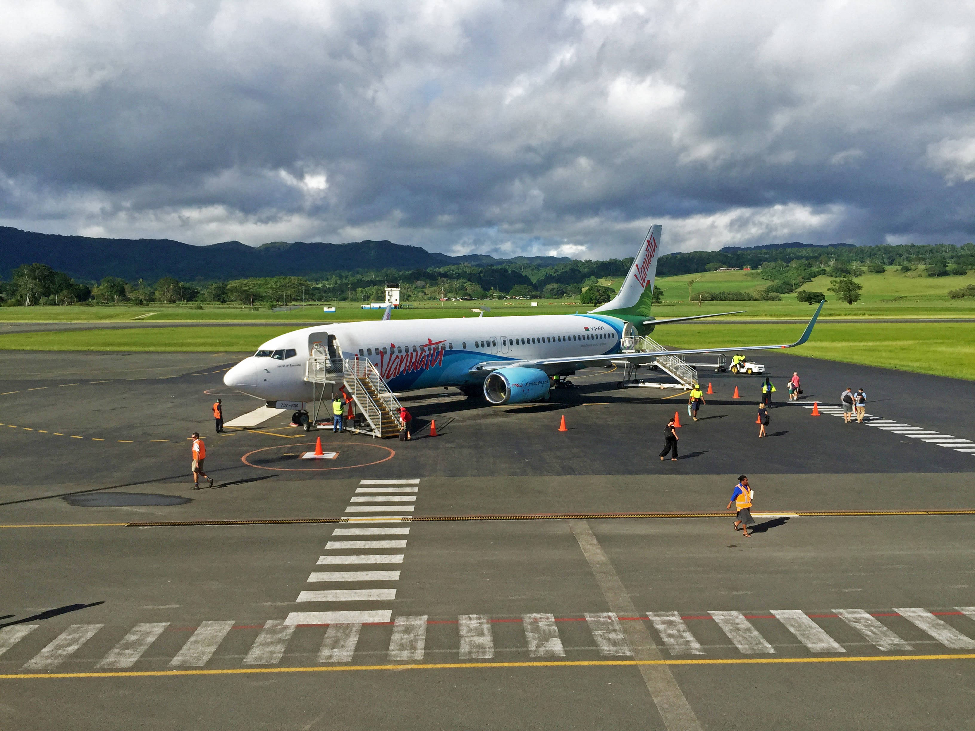 Vanuatu 桌布 壁紙 — 一架維珍澳洲航空飛機停在停機坪上，背景是戲劇性的暴風雲，附近有地勤人員和乘客。