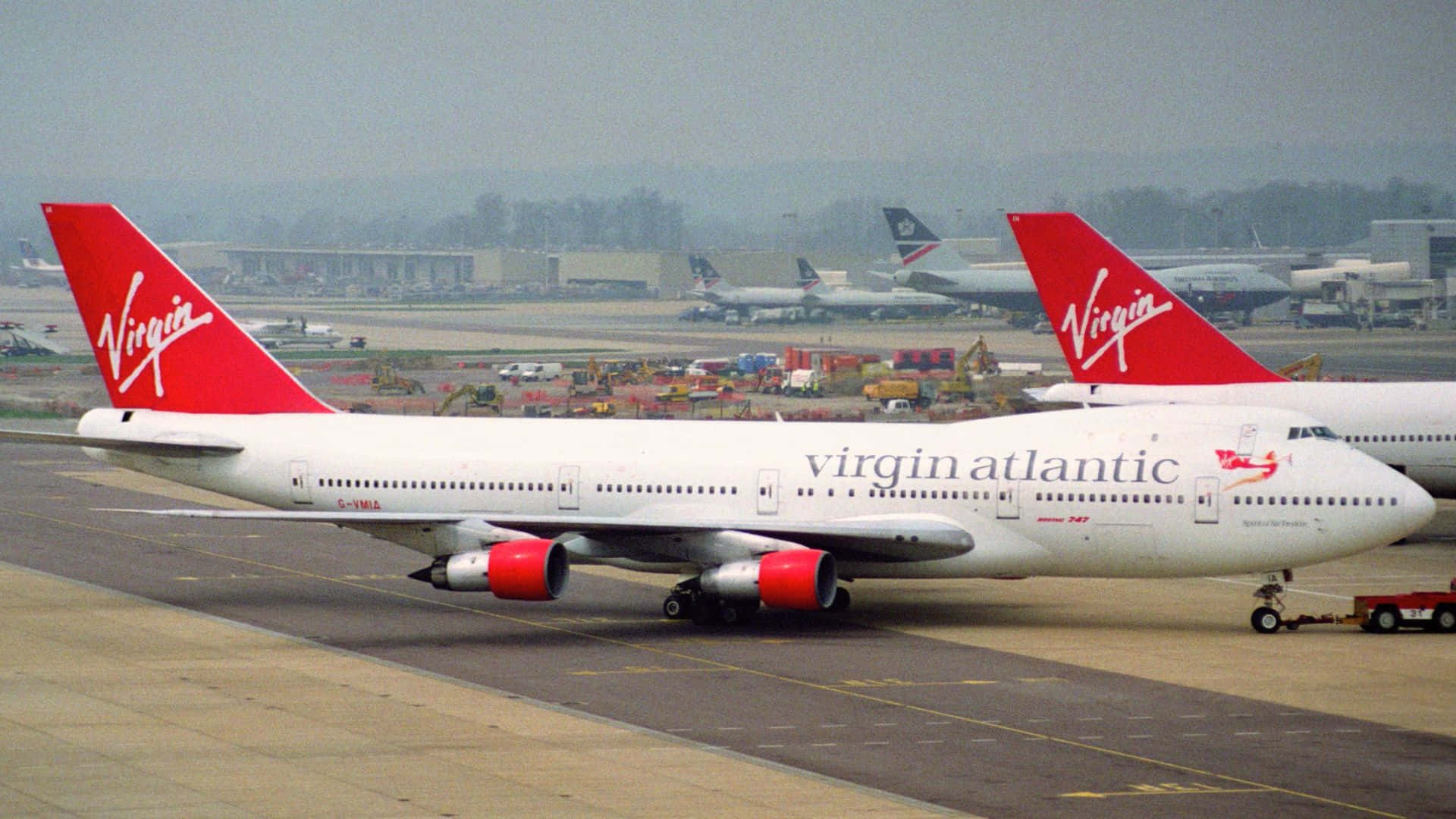 1920x1080 Jumbo Jets 桌布 壁紙 — 一架維珍航空波音747在霧濛濛的機場停機坪滑行，背景有其他飛機和地面設備。