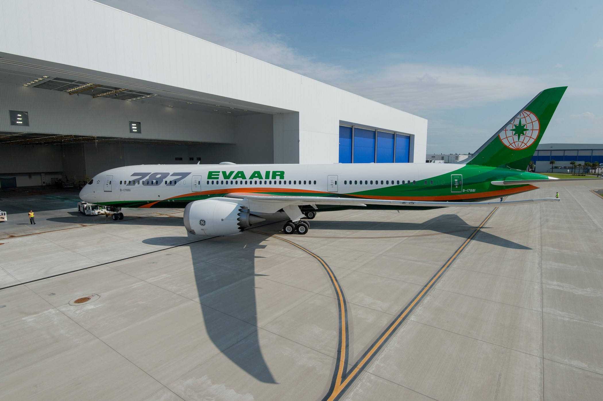 Airport 桌布 壁紙 — 一架線條流暢的EVA AIR波音787夢幻客機，採用綠色、橙色和白色塗裝，停在現代化機庫外的停機坪上，天空清澈湛藍。