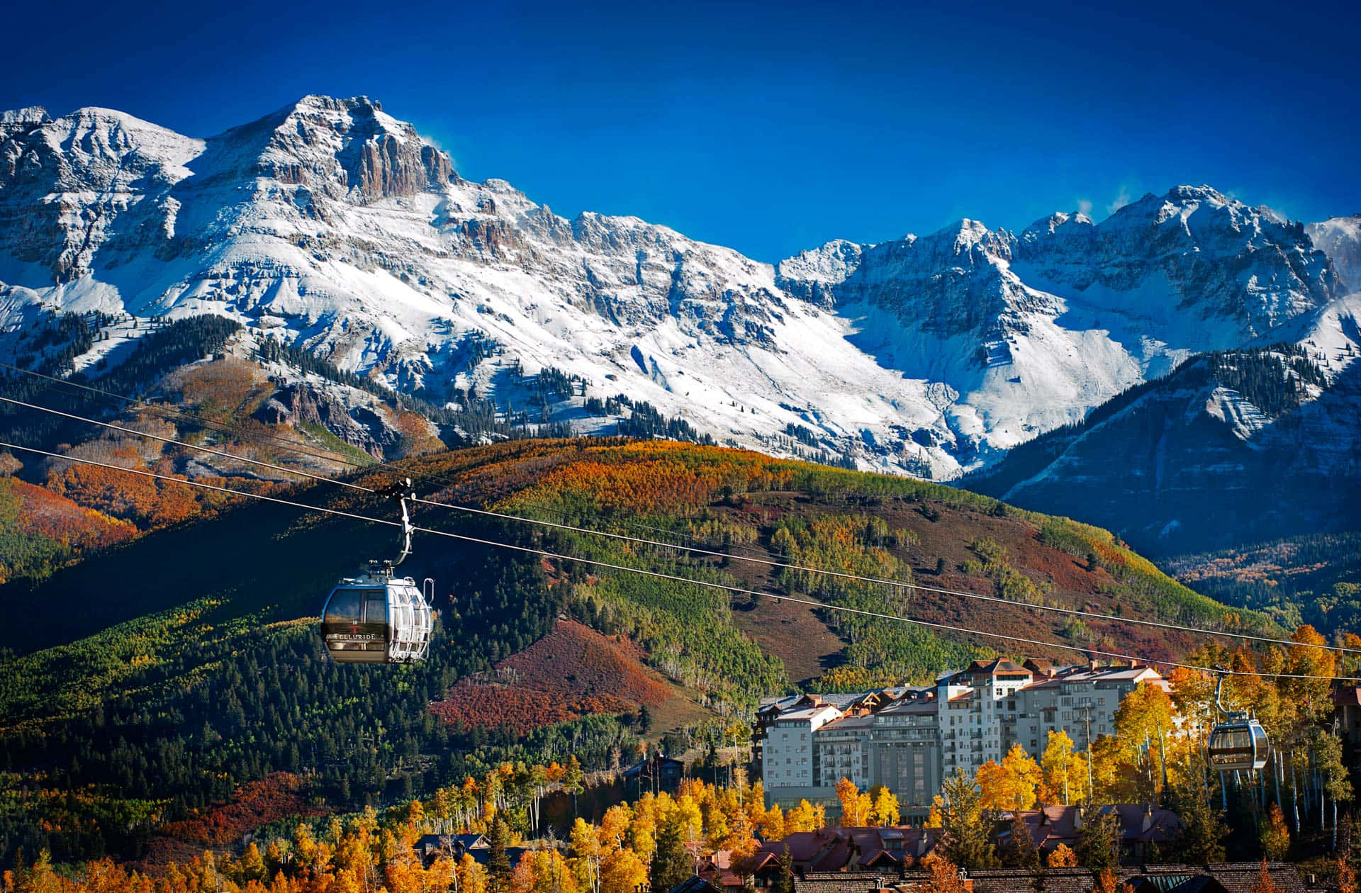 Colorado Mountain 桌布 壁紙 — 一架纜車在晴朗藍天下，滑過一個充滿活力的秋季山谷，山谷中有積雪山峰和一個度假小鎮。