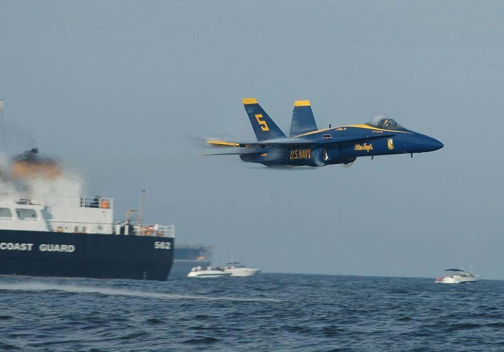 US Navy 桌布 壁紙 — 一架美國海軍藍天使特技飛行隊的噴射機低飛過波濤洶湧的海面，下方有海岸警衛隊船隻和快艇。