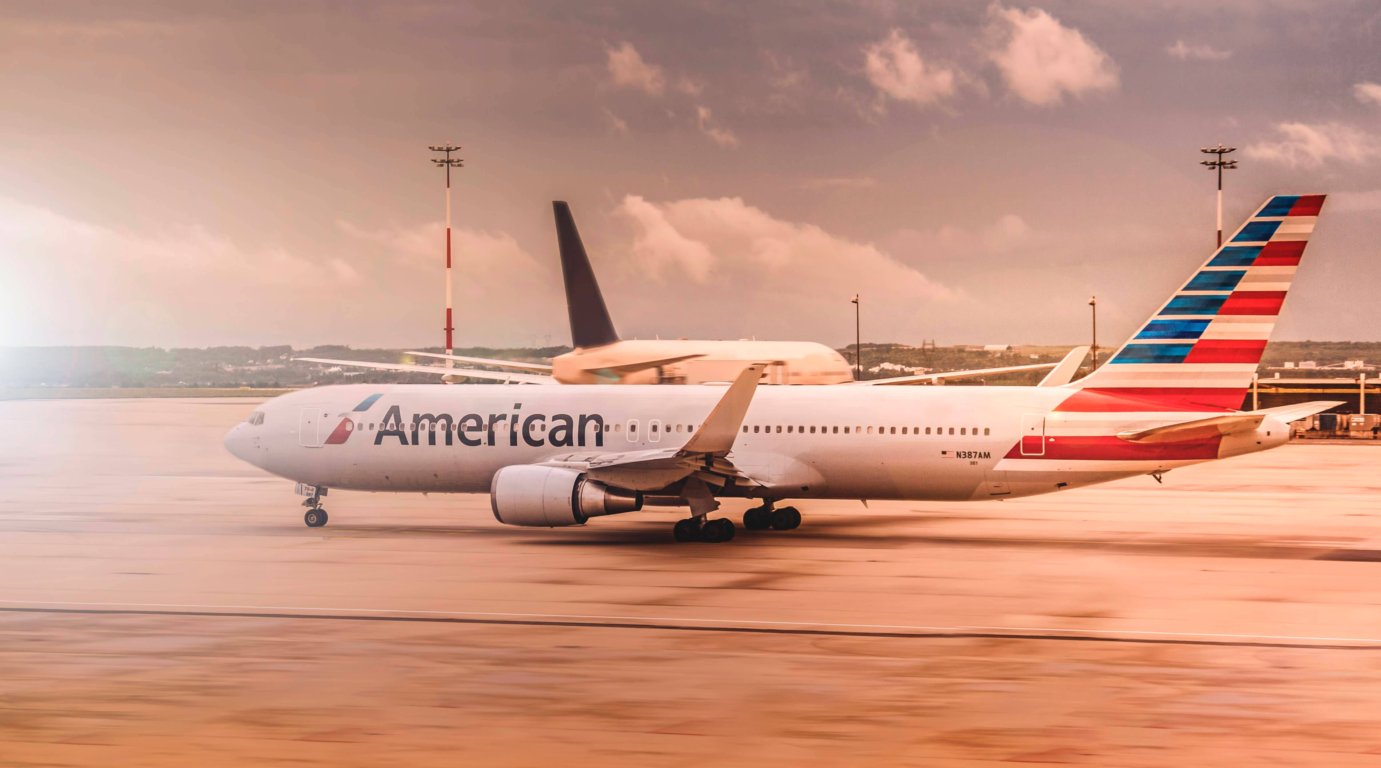 Airlines 桌布 壁紙 — 一架美國航空噴射機在陽光普照的停機坪上滑行，天空多雲，遠處有機場燈光。