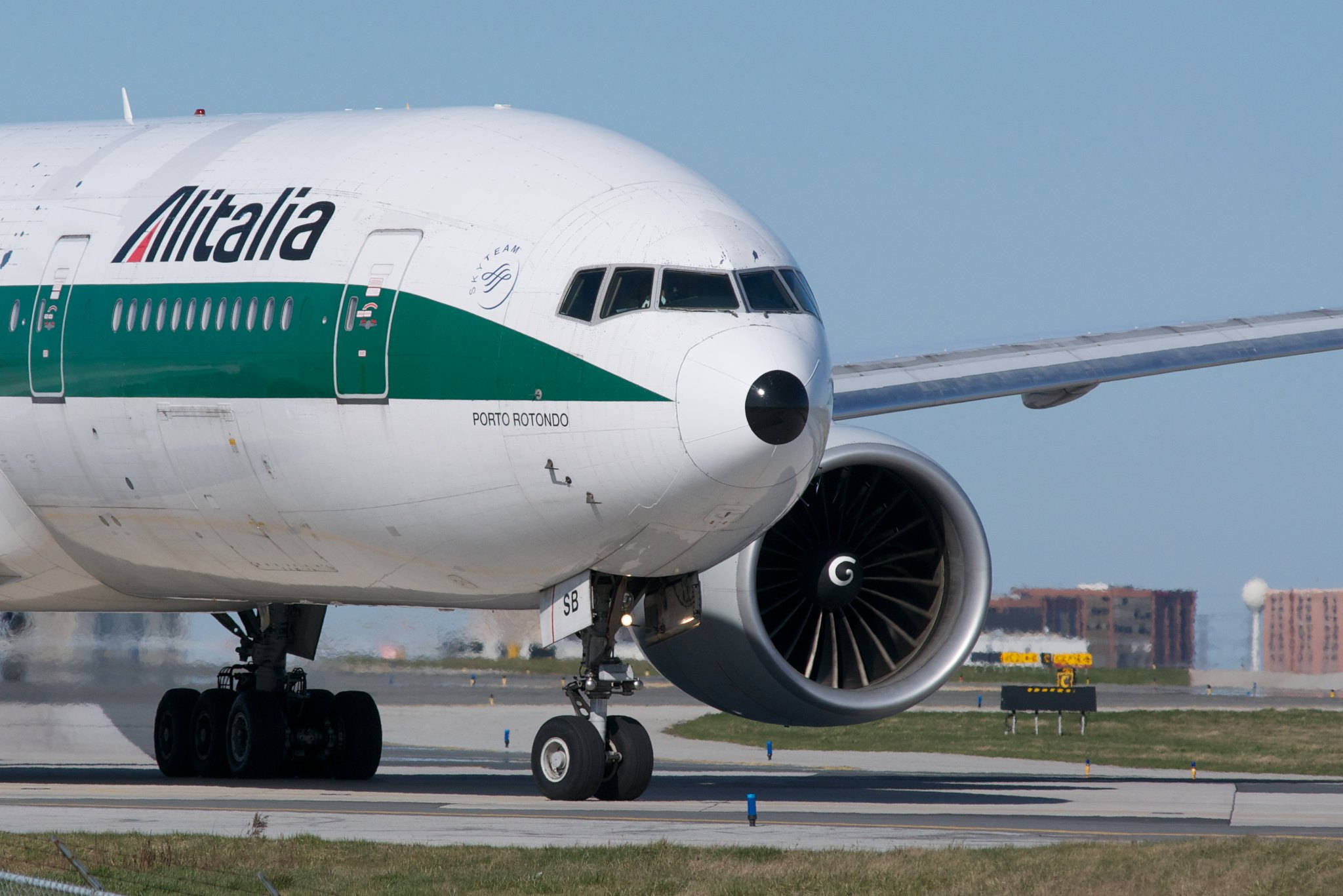 飛機 桌布 壁紙 — 一架義大利航空的波音777在陽光明媚的機場跑道上滑行，天空清澈湛藍，遠處有建築物。
