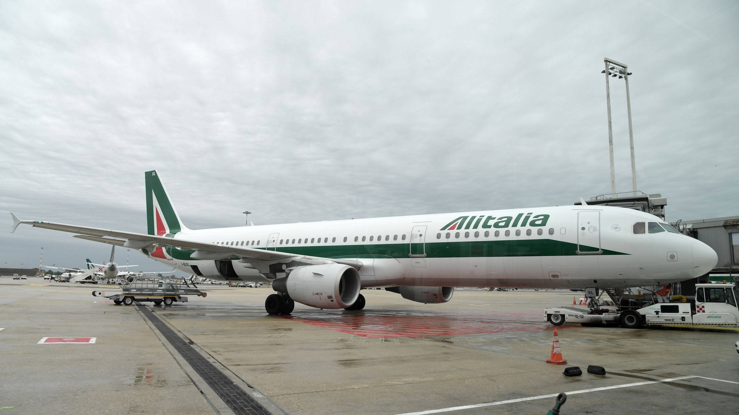 飛機 桌布 壁紙 — 一架義大利航空飛機停在機場閘口，濕滑的停機坪上，天空多雲，附近有地勤人員和設備。