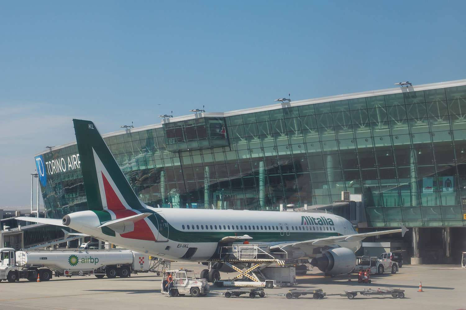  壁紙 — 一架義大利航空飛機停泊在杜林機場航廈，天空晴朗蔚藍，附近有地勤人員和服務車輛。