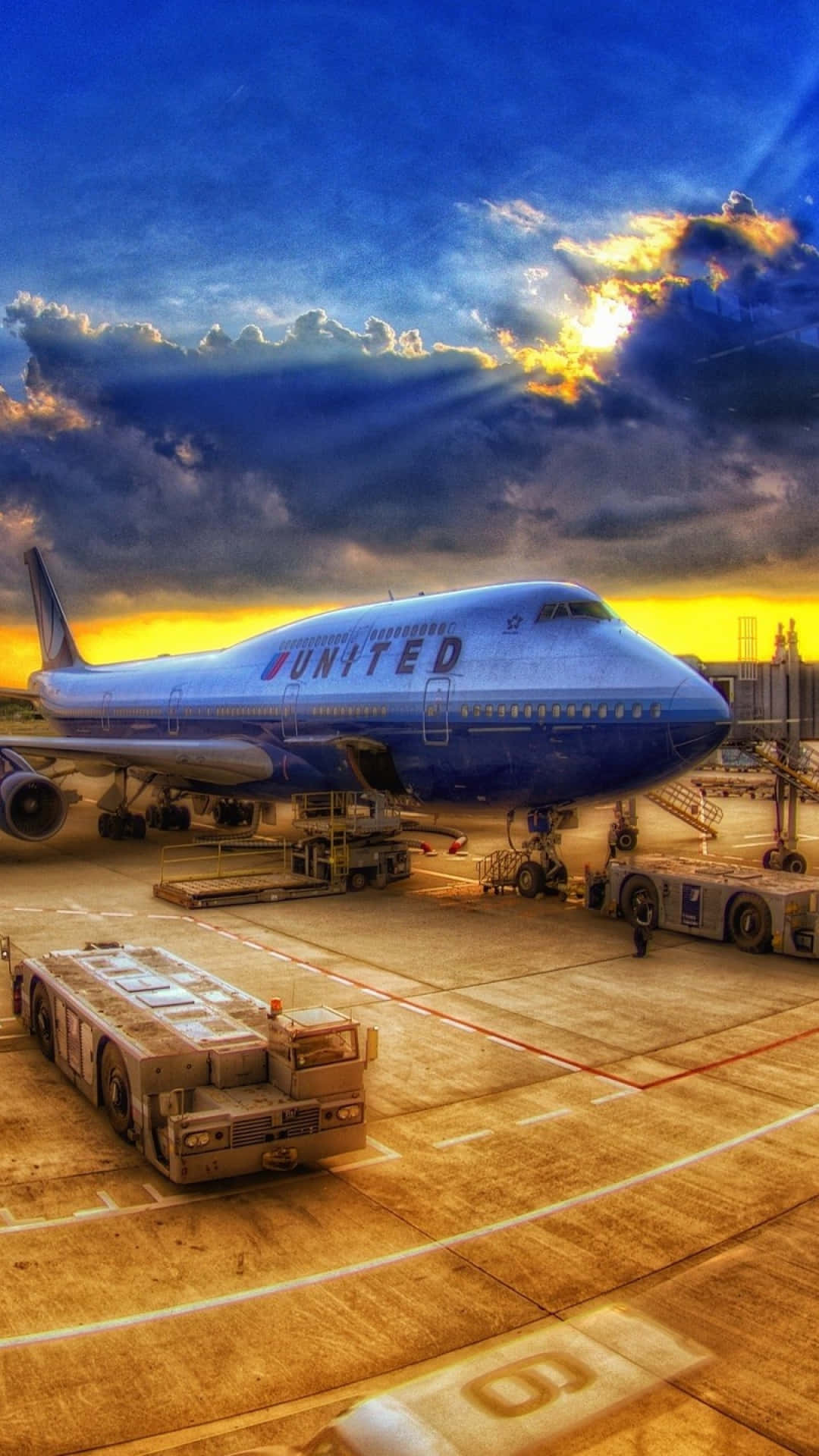 Plane 桌布 壁紙 — 一架聯合航空珍寶客機停在機場閘口，背景是戲劇性的日落天空，陽光穿透雲層射出耀眼的光芒。