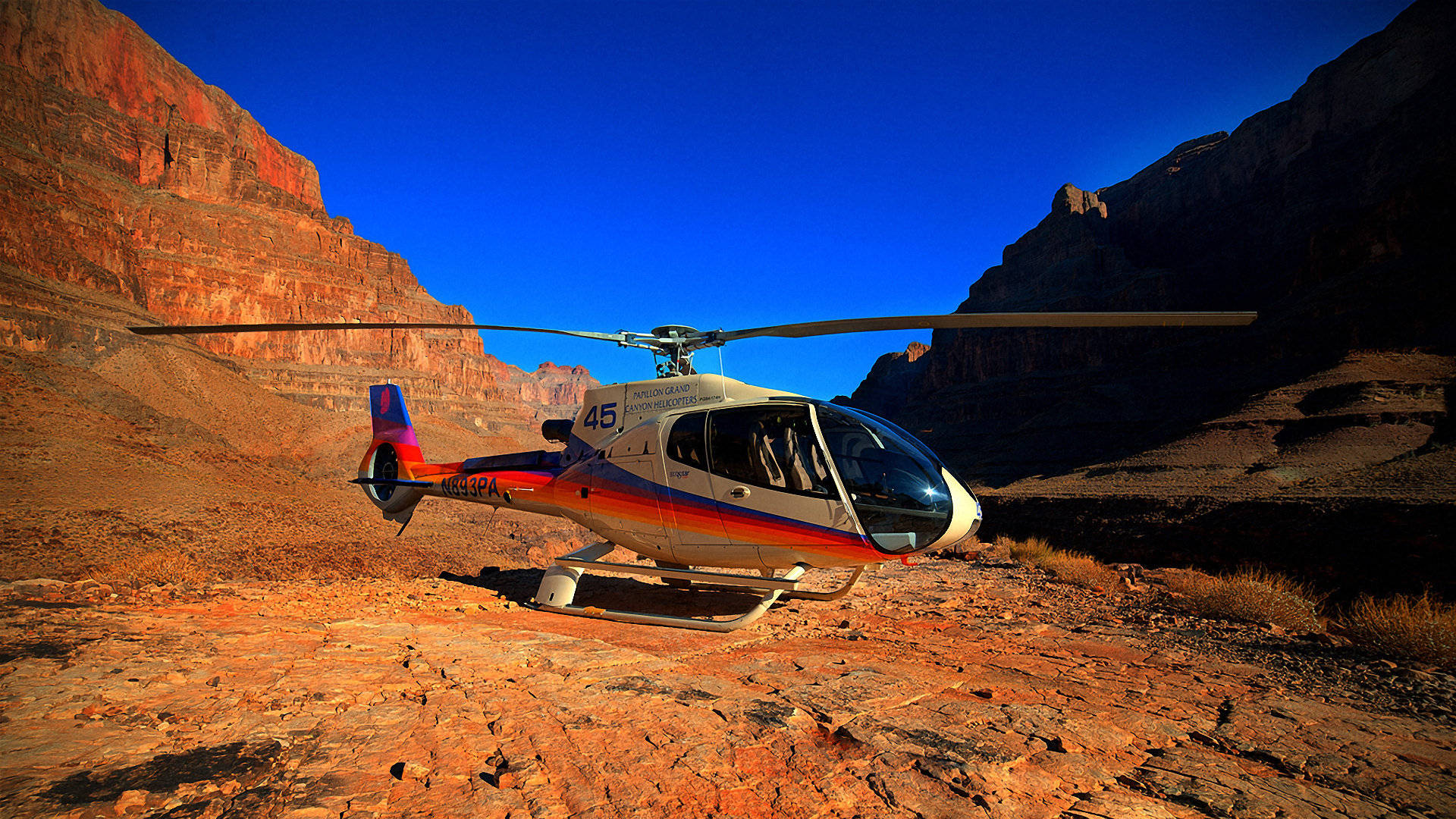 helicopter 4K 桌布 壁紙 — 一架色彩繽紛的直升機停泊在崎嶇的紅色岩石地形上，背景是廣闊峽谷中鮮明的藍天。