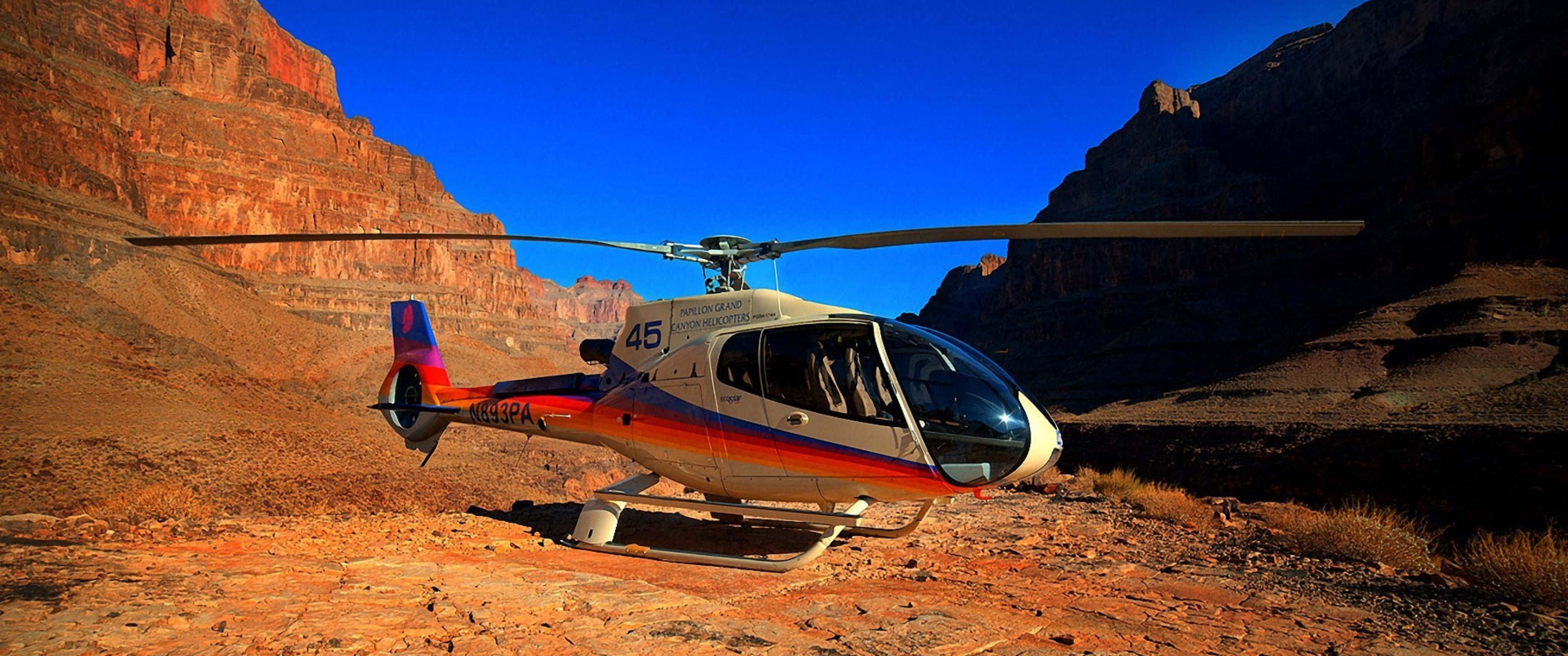 3440x1440p Helicopters 桌布 壁紙 — 一架色彩繽紛的直升機停泊在崎嶇的紅色岩石地形上，背景是廣闊峽谷中清澈的藍天。