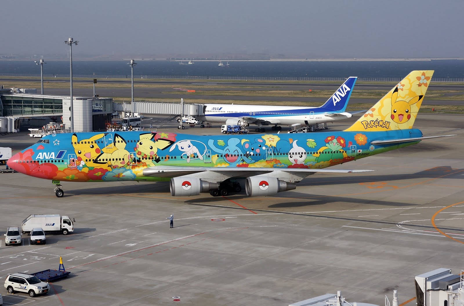 飛機 桌布 壁紙 — 一架色彩鮮豔的ANA寶可夢主題飛機停在機場停機坪上，背景有另一架飛機和海洋。