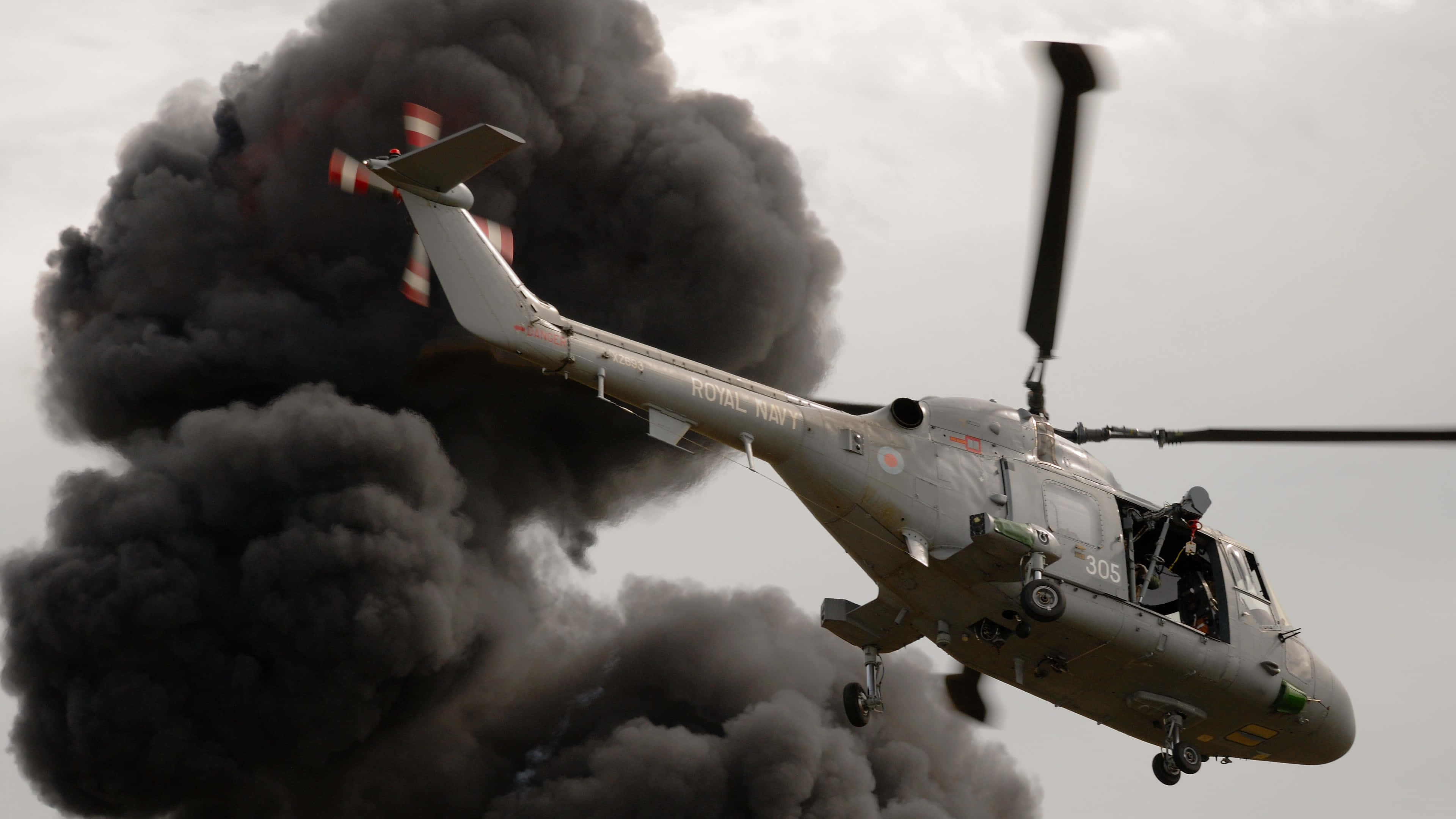 4K Helicopters 桌布 壁紙 — 一架英國皇家海軍直升機在灰色天空背景下飛過濃厚黑煙。