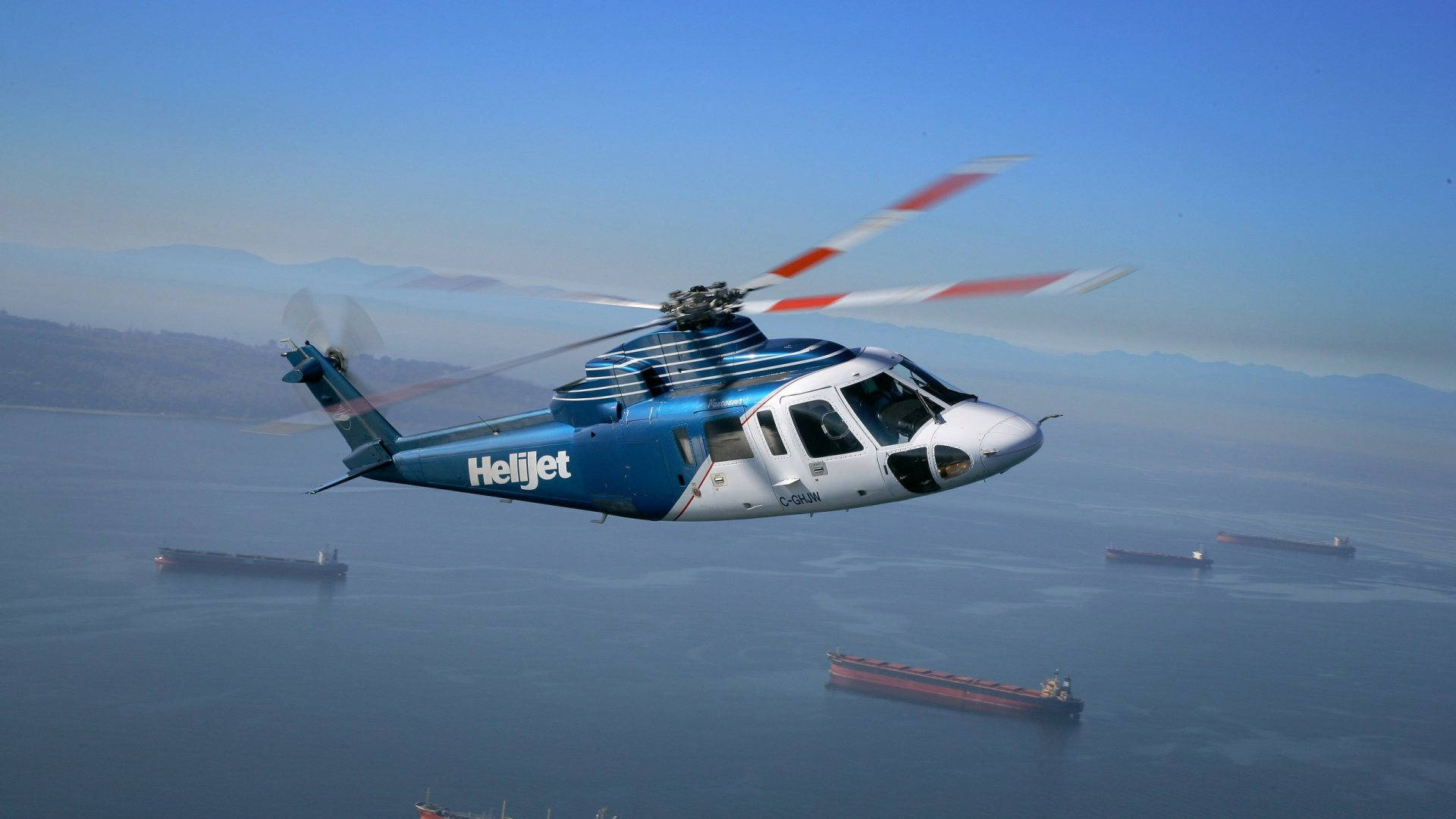 helicopter 4K 桌布 壁紙 — 一架藍白色的Helijet直升機低飛過平靜的海面，下方有貨船，遠處有朦朧的山巒。