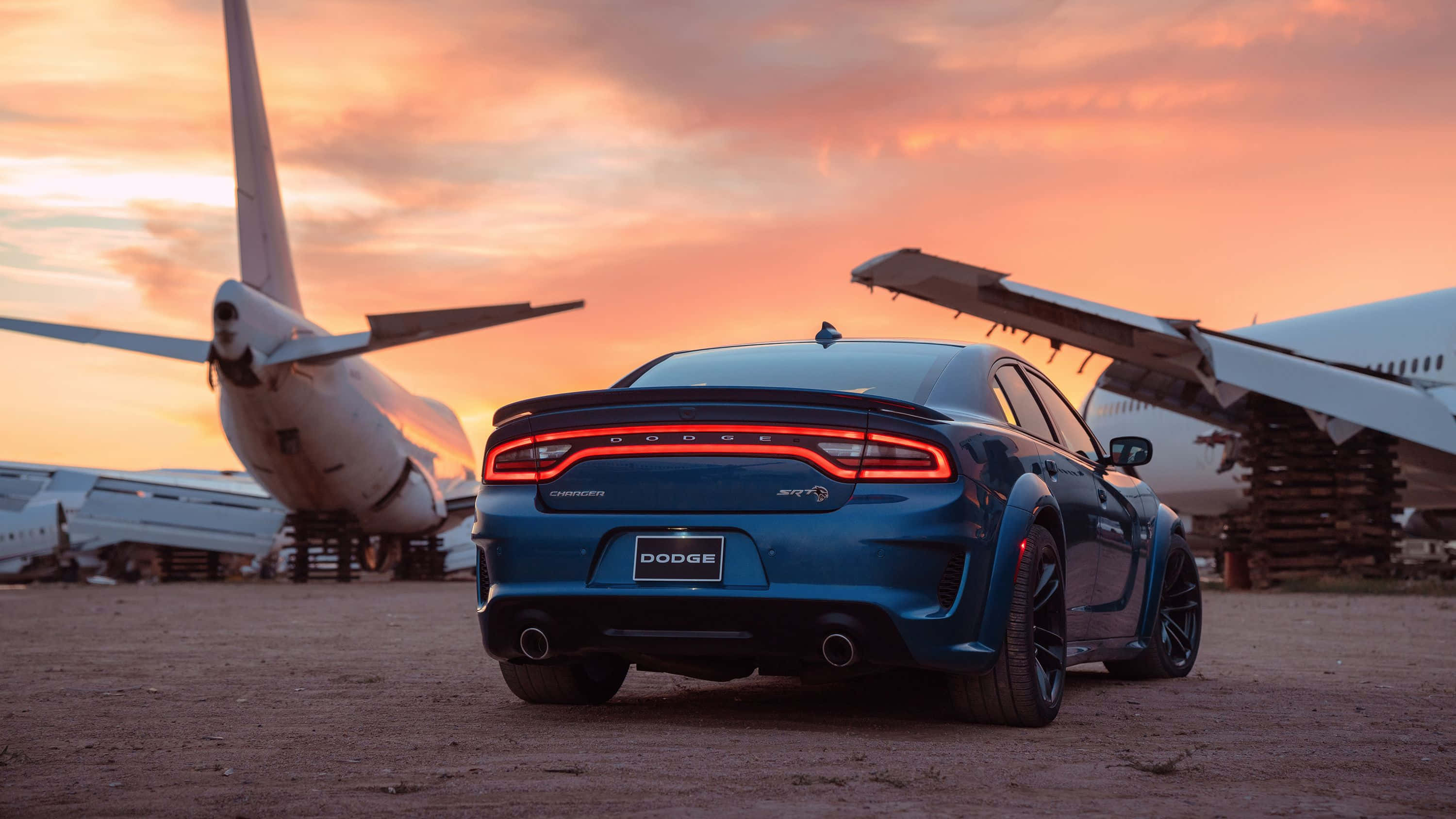 Dodge Charger 桌布 壁紙 — 一架藍色Dodge Charger SRT停放在兩架退役飛機之間，背景是熾熱的日落天空。