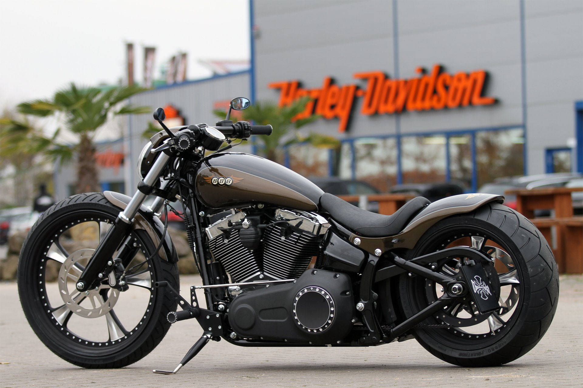 Bobber 桌布 壁紙 — 一架訂製消光黑色哈雷機車停放在經銷商前，周圍有棕櫚樹和陰天。