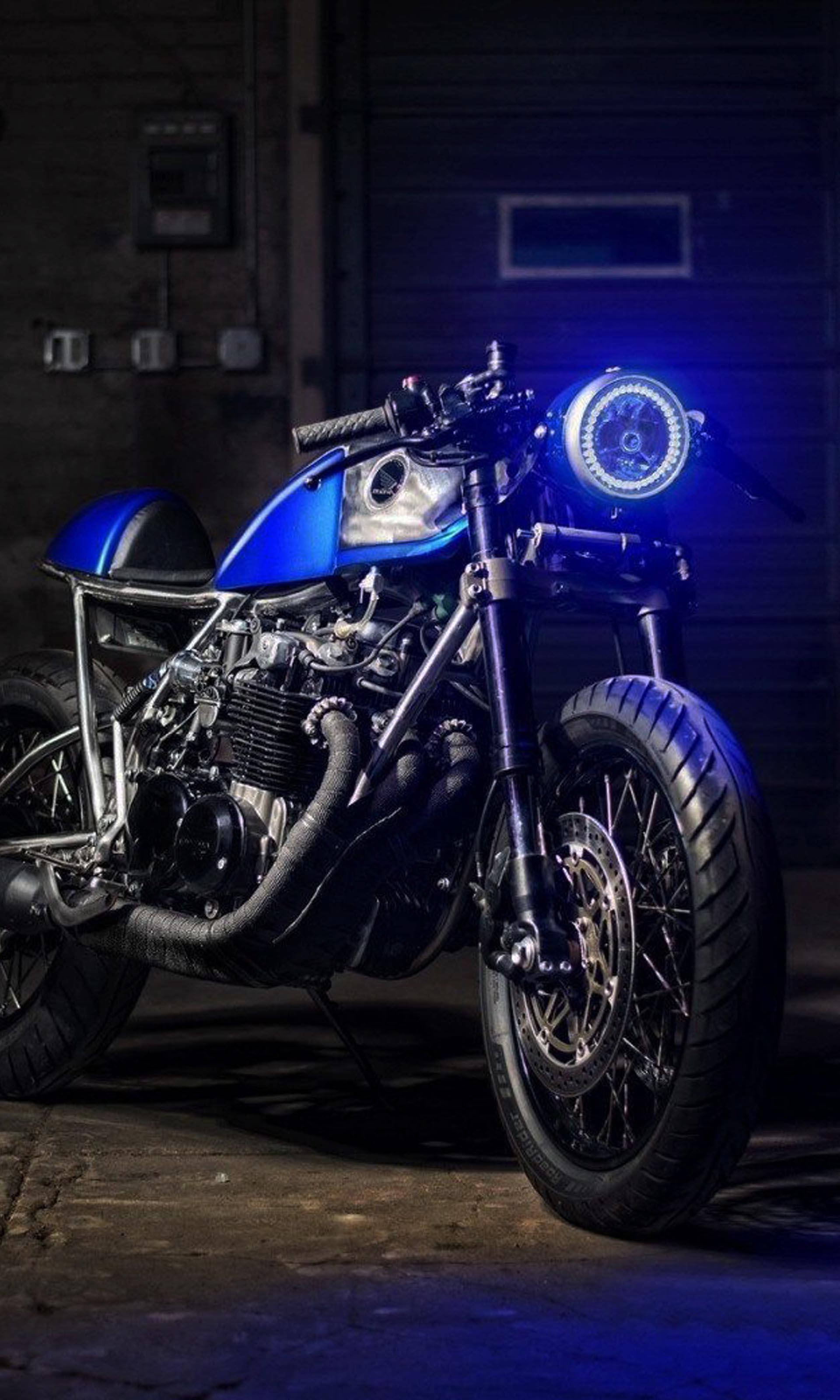 4K bike 桌布 壁紙 — 一架訂製的藍色咖啡賽車摩托車，帶有發光的頭燈，停在光線昏暗的車庫中，在混凝土地板上投射出冷藍色的光芒。