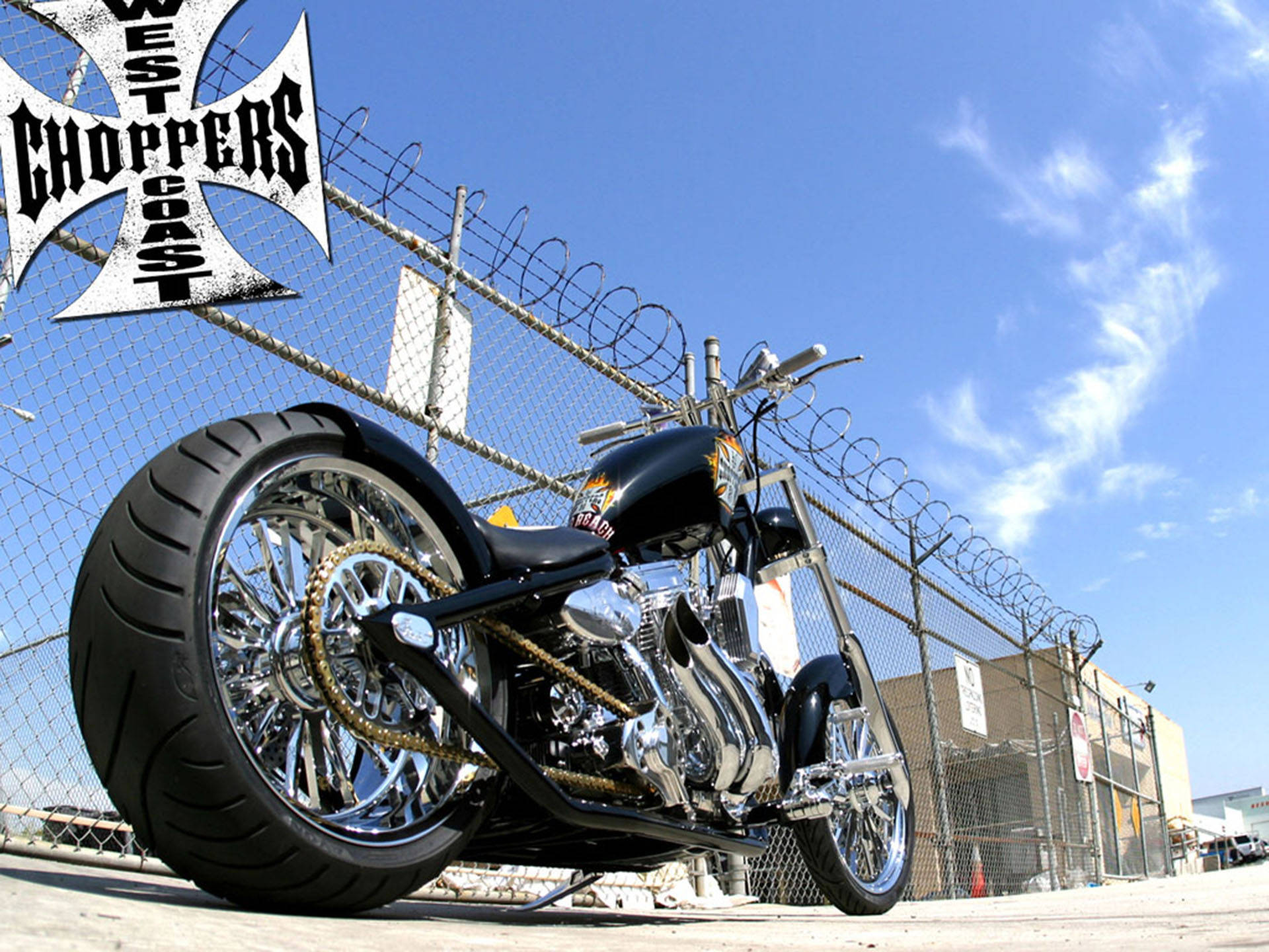 Choppers 桌布 壁紙 — 一架訂製黑色chopper機車，帶有鍍鉻細節和大前輪胎，停在鏈條圍欄前，背景是晴朗的藍天。