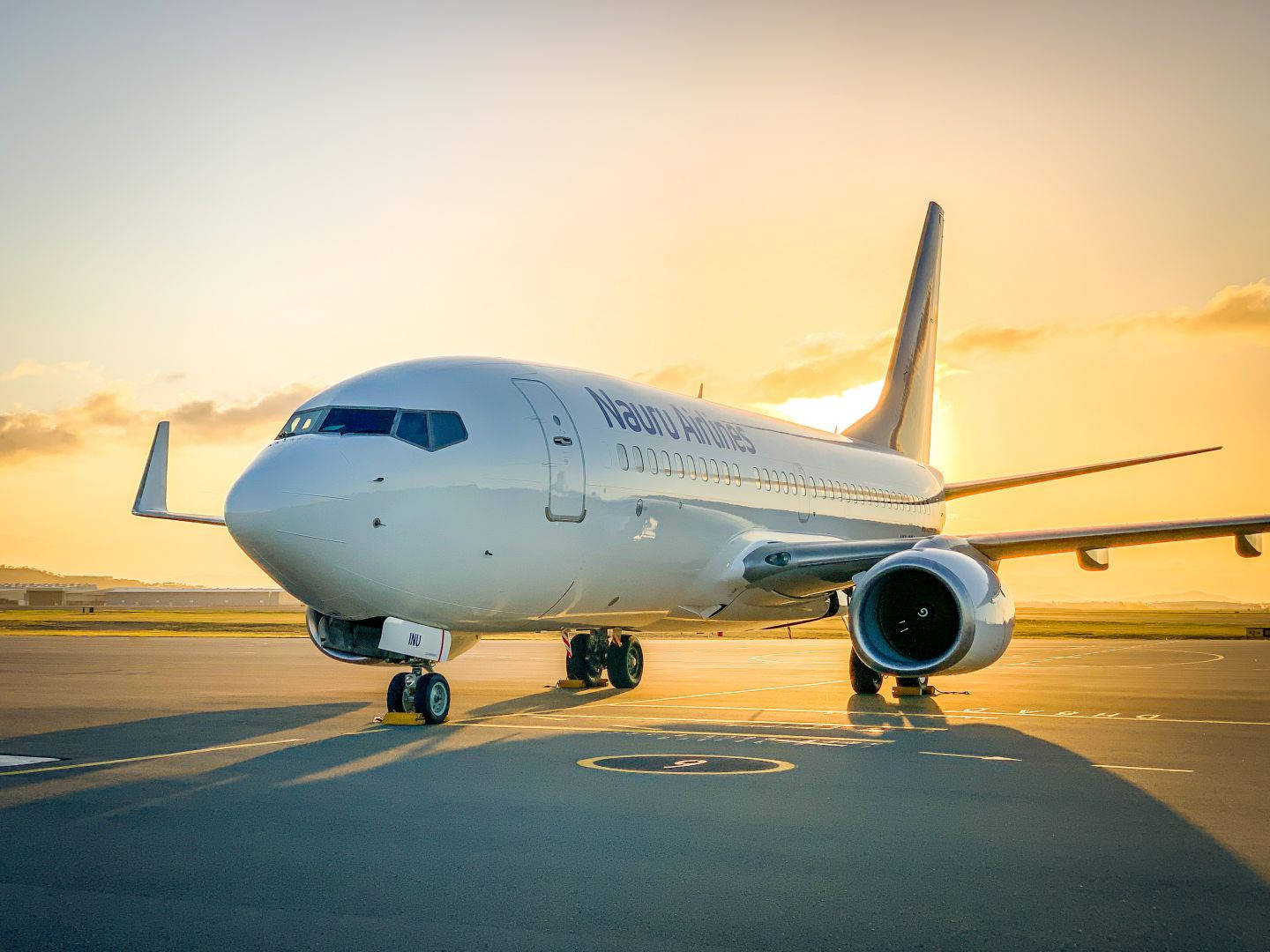 飛機 桌布 壁紙 — 一架諾魯航空客機停在機場停機坪上，沐浴在金色的夕陽中，投下長長的影子。