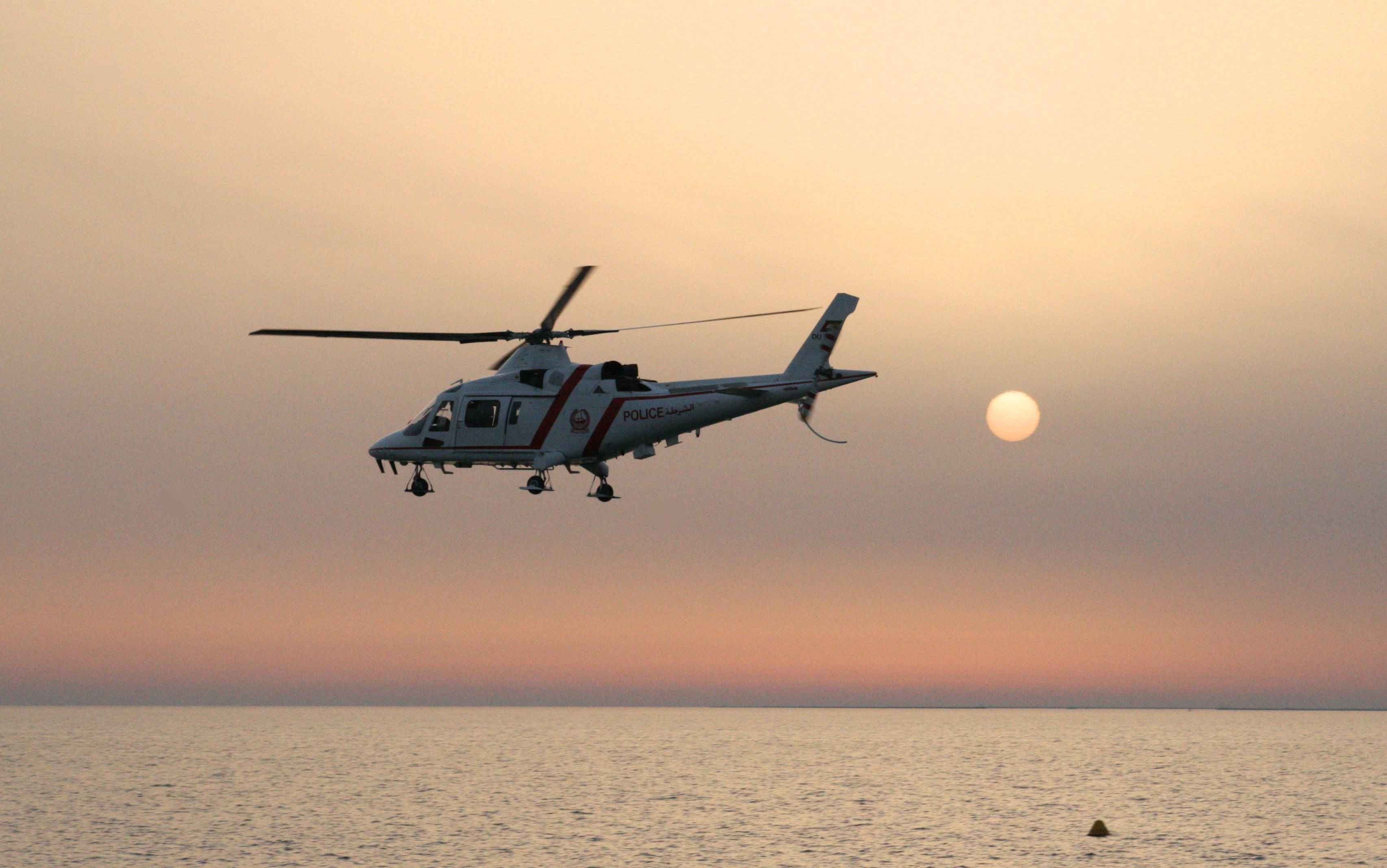 helicopter 4K 桌布 壁紙 — 一架警用直升機在平靜的海面上盤旋，夕陽在柔和的橙粉色的天空中緩緩落下。