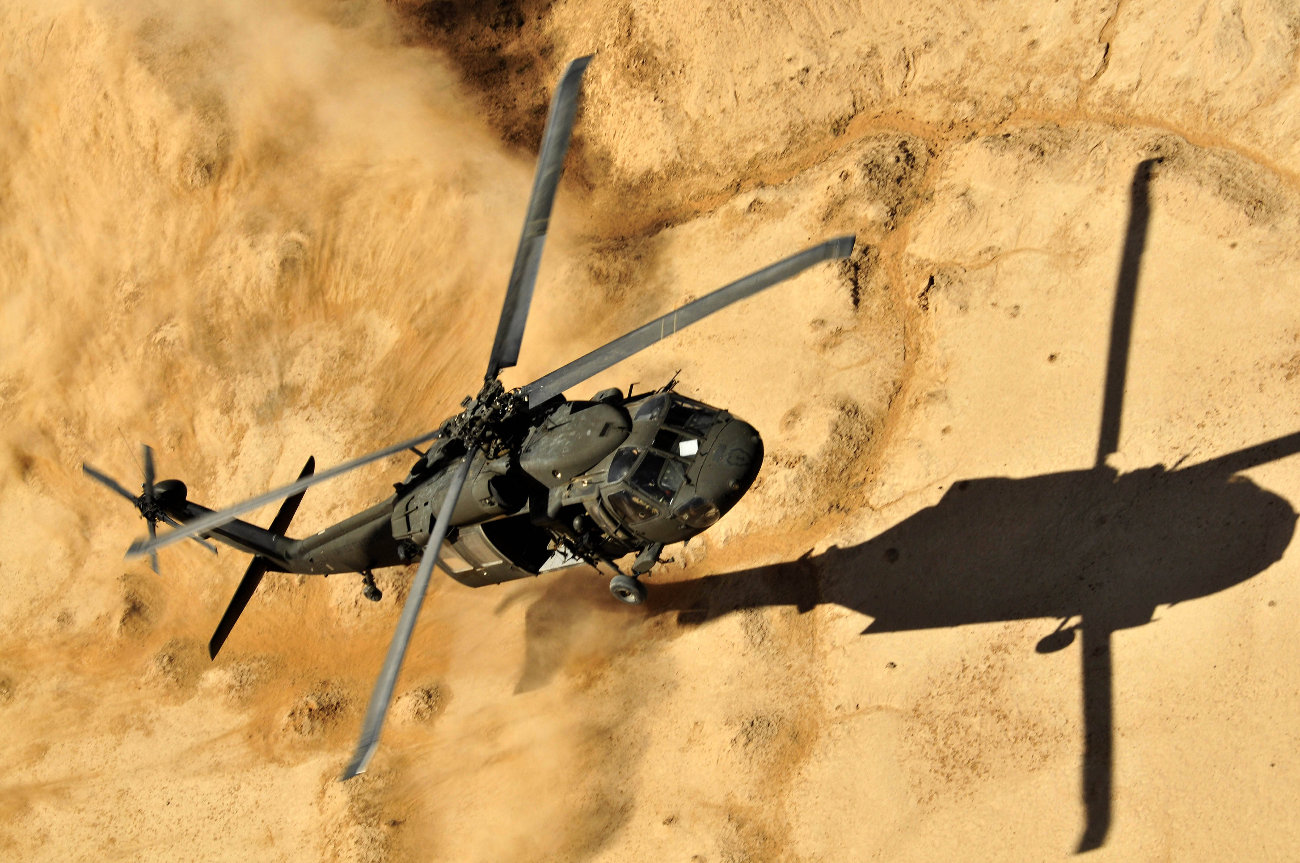 Helicopter 桌布 壁紙 — 一架軍用直升機低空盤旋在乾旱的沙漠地形上，揚起沙塵，其影子在沙丘上拉得很長。