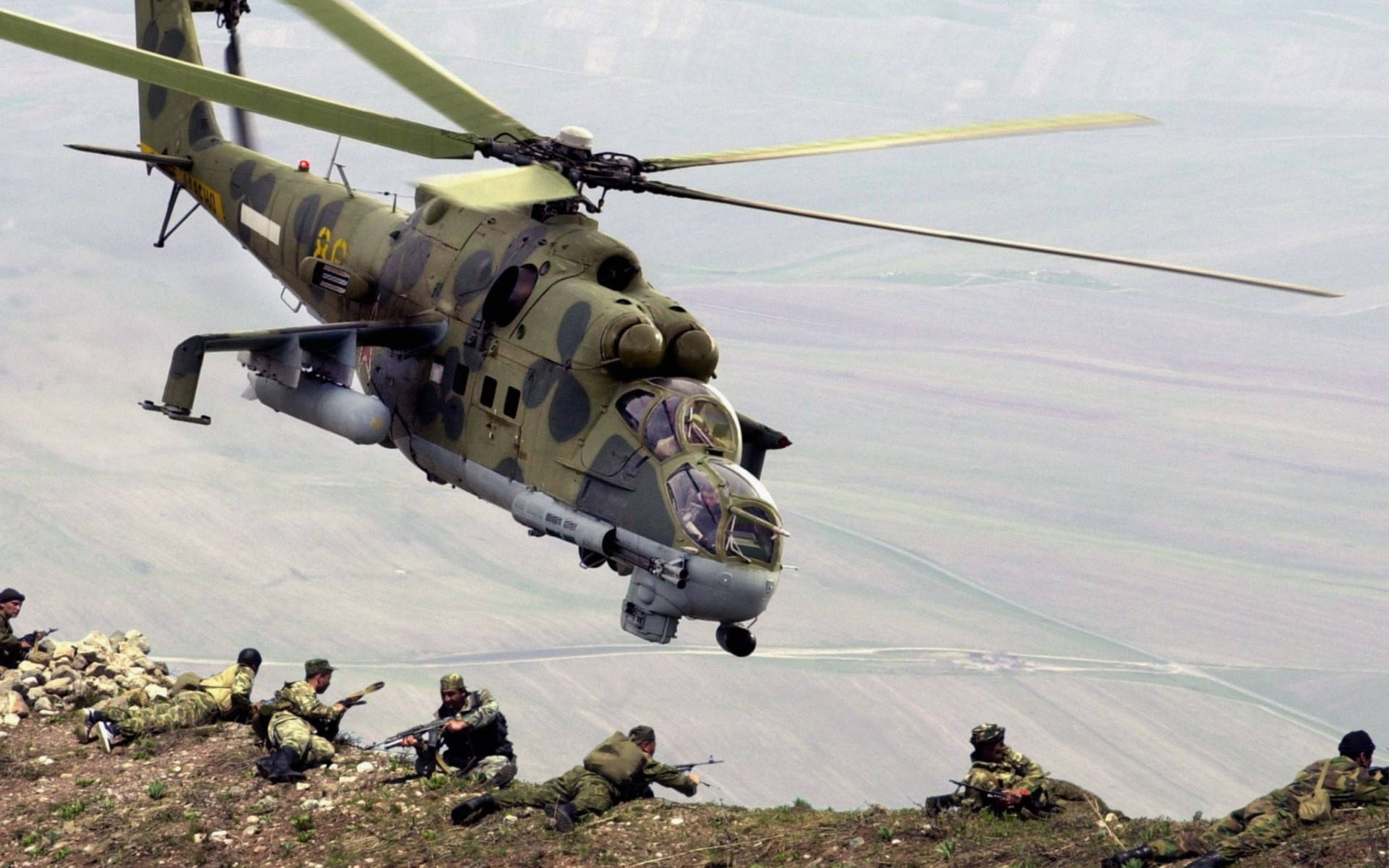 Helicopter 桌布 壁紙 — 一架軍用直升機低空盤旋在身穿迷彩裝備的士兵上方，這些士兵位於岩石山坡上，俯瞰廣闊的山谷。