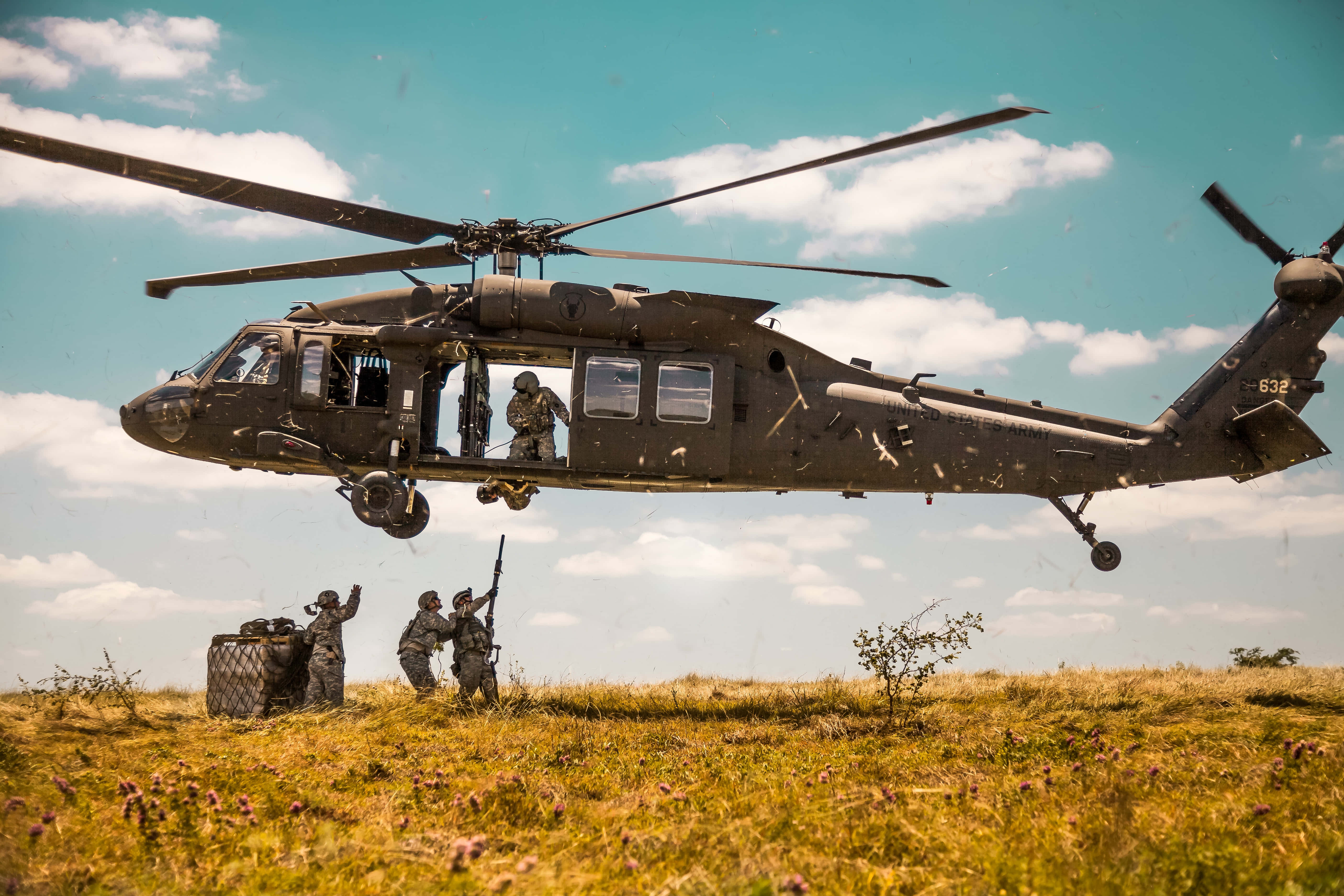Helicopter 桌布 壁紙 — 一架軍用直升機低空盤旋在陽光照耀的草地上，士兵們在晴朗藍天和零星白雲下準備部署。