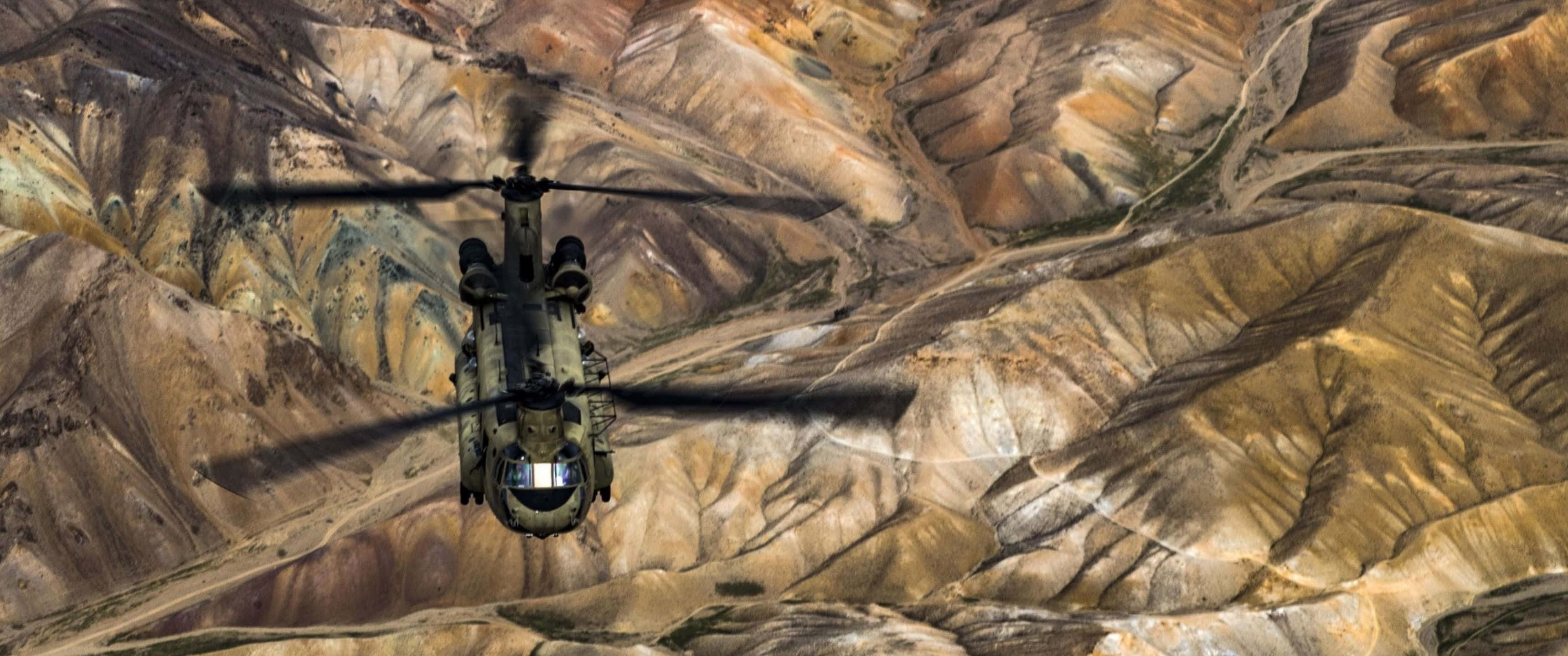 3440x1440p Helicopters 桌布 壁紙 — 一架軍用直升機低飛過一個崎嶇的峽谷，峽谷呈現層層泥土色調和尖銳的山脊。