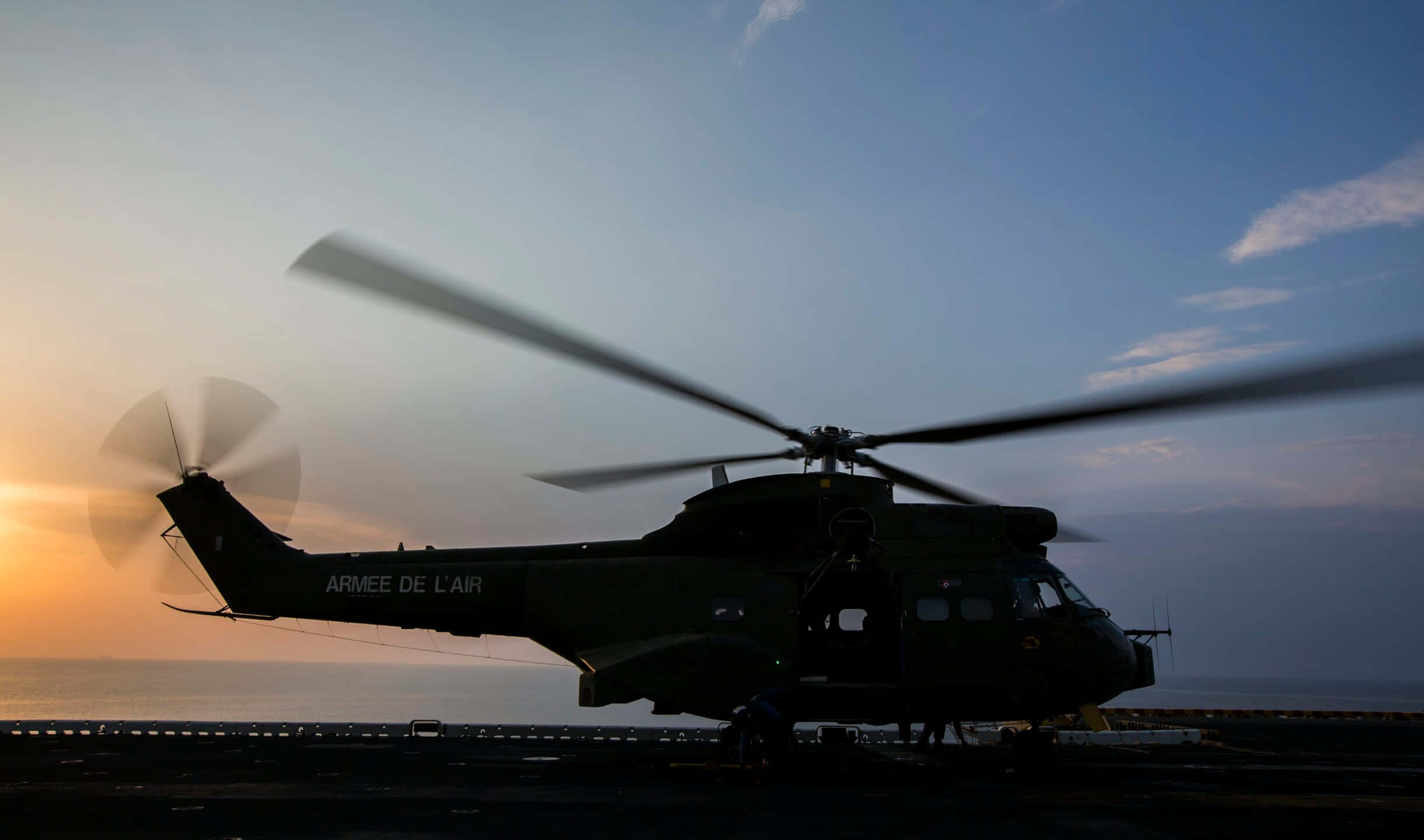2440x1440 Helicopters 桌布 壁紙 — 一架軍用直升機在夕陽天空的襯托下形成剪影，盤旋在船隻甲板上方。