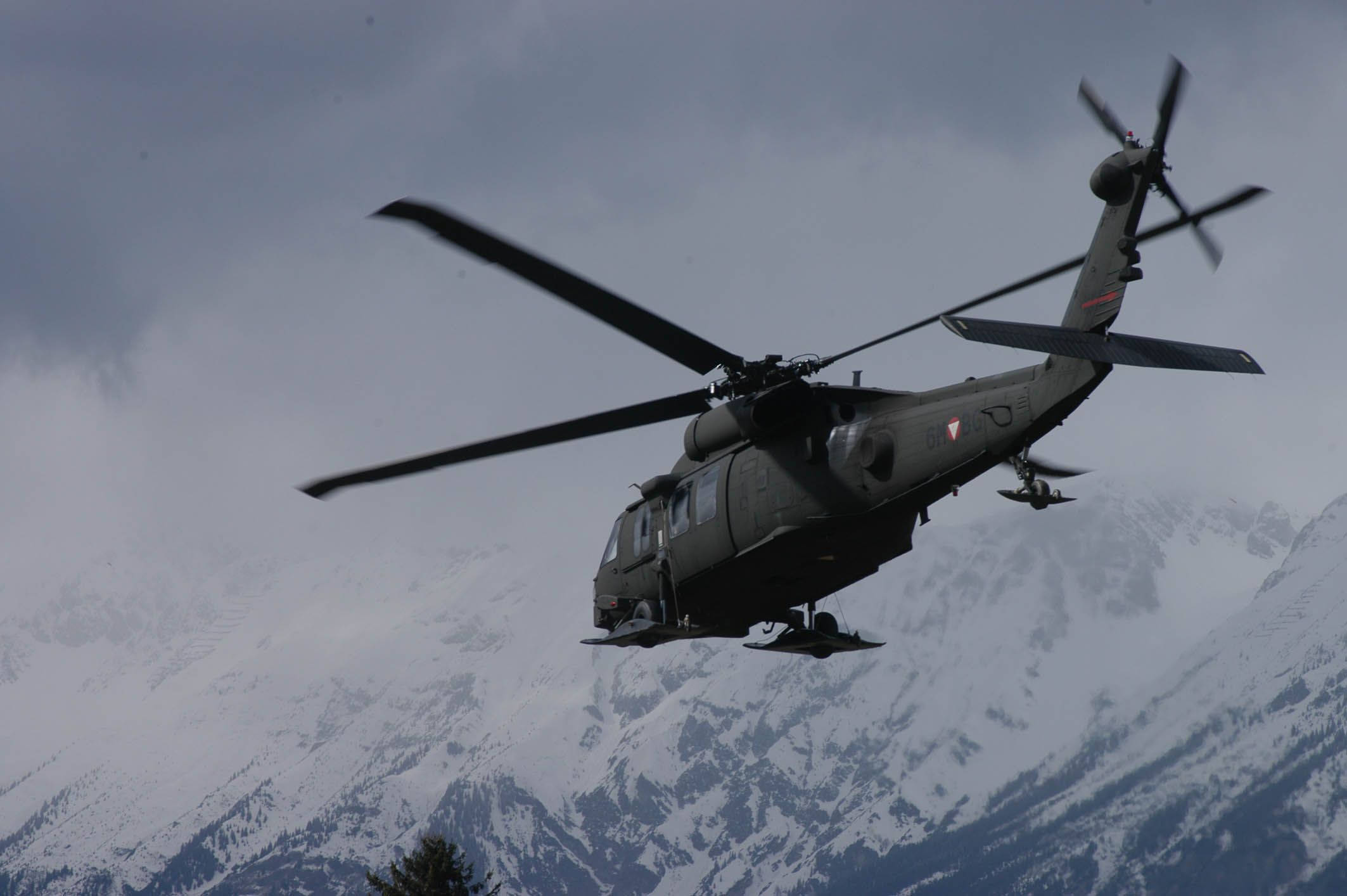 Helicopter 桌布 壁紙 — 一架軍用直升機在多雲的天空下低飛越過積雪的山峰。