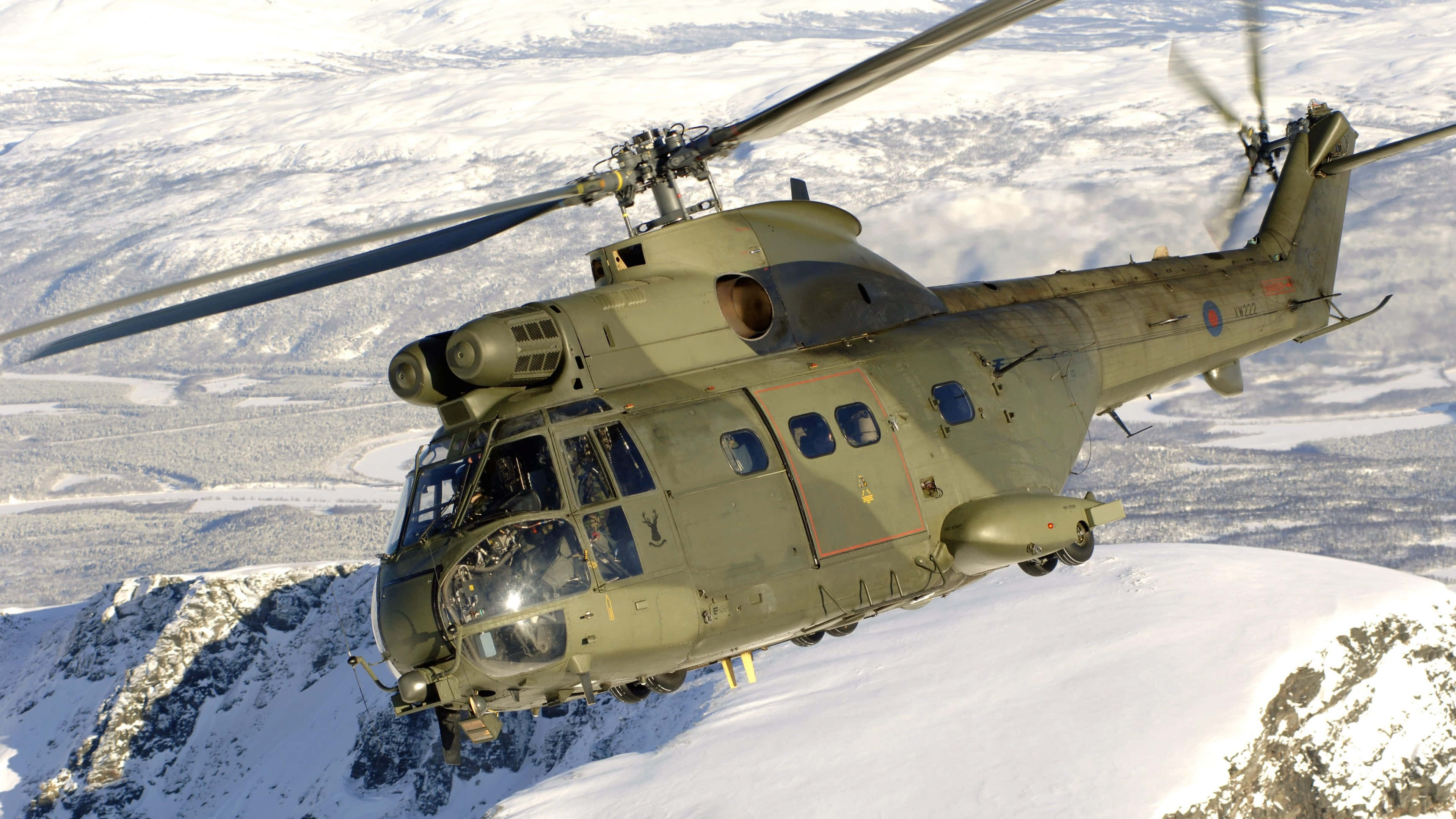 4K Helicopters 桌布 壁紙 — 一架軍用直升機在明亮的日光下低飛越過積雪覆蓋的山脈。