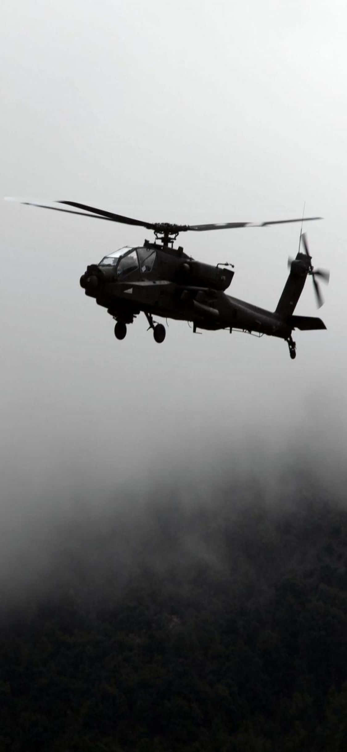 iPhone Xs Helicopters 桌布 壁紙 — 一架軍用直升機在灰色天空的襯托下呈現剪影，盤旋在濃霧籠罩的茂密森林上方。