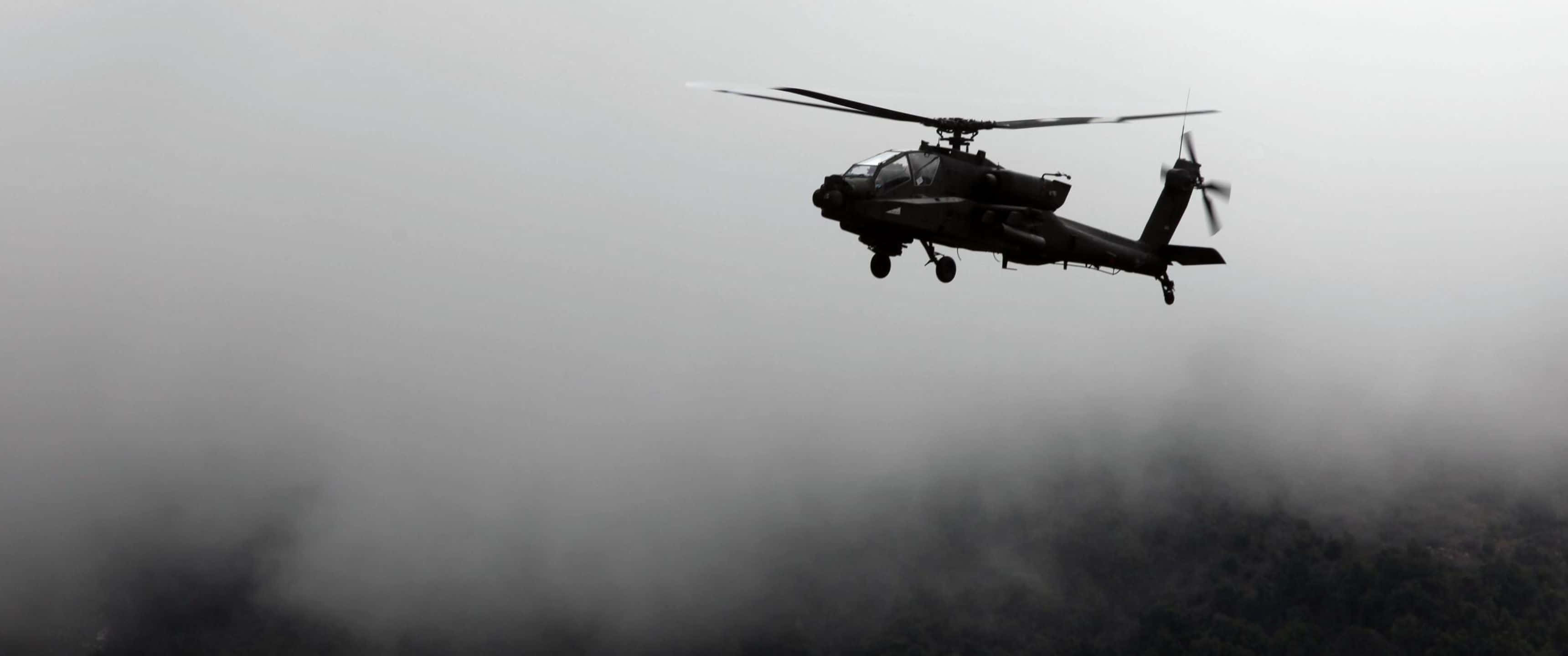 3440x1440p Helicopters 桌布 壁紙 — 一架軍用直升機在陰天天空的襯托下，盤旋在霧氣籠罩的森林景觀上方。
