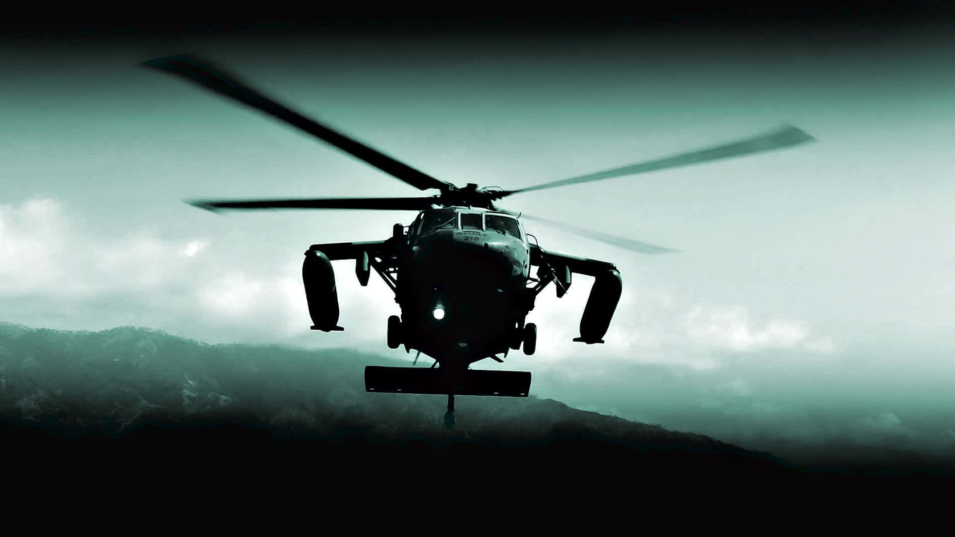 Helicopter 桌布 壁紙 — 一架軍用直升機在陰雲密佈的天空中呈現剪影，在戲劇性的低光場景中盤旋於霧氣繚繞的山巒之上。