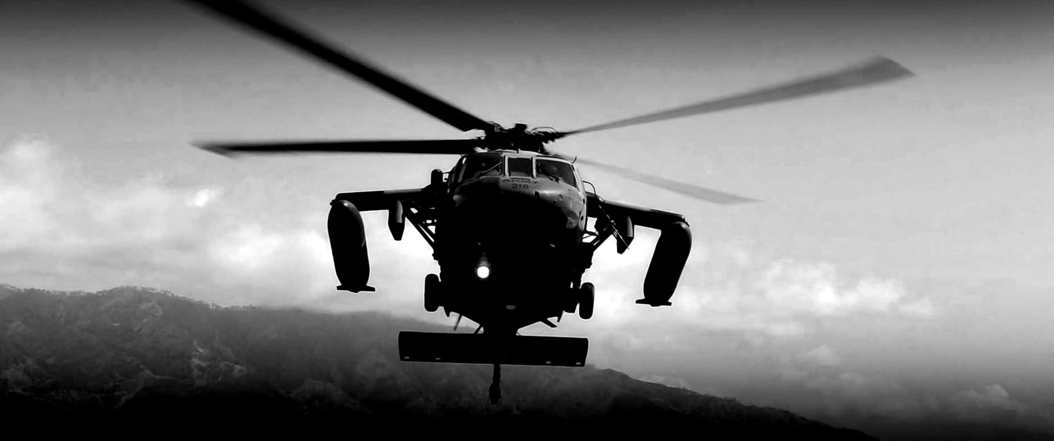 3440x1440p Helicopters 桌布 壁紙 — 一架軍用直升機在陰雲密布的天空中呈現剪影，盤旋在崎嶇的山地地形之上，以鮮明的黑白呈現。