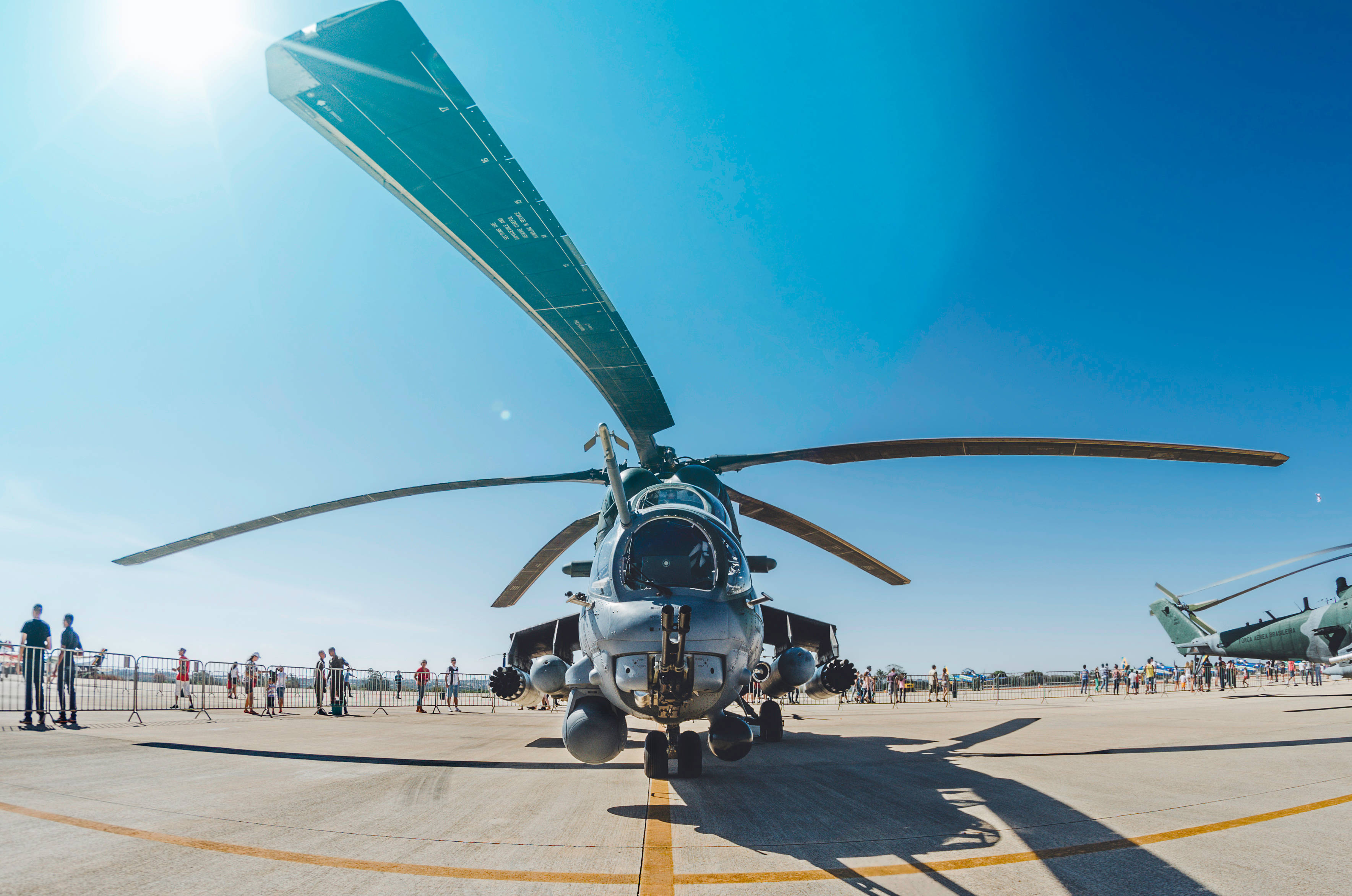 Helicopter 桌布 壁紙 — 一架軍用直升機在陽光明媚的機場上空盤旋，人們在晴朗的藍天下從障礙物後觀察。