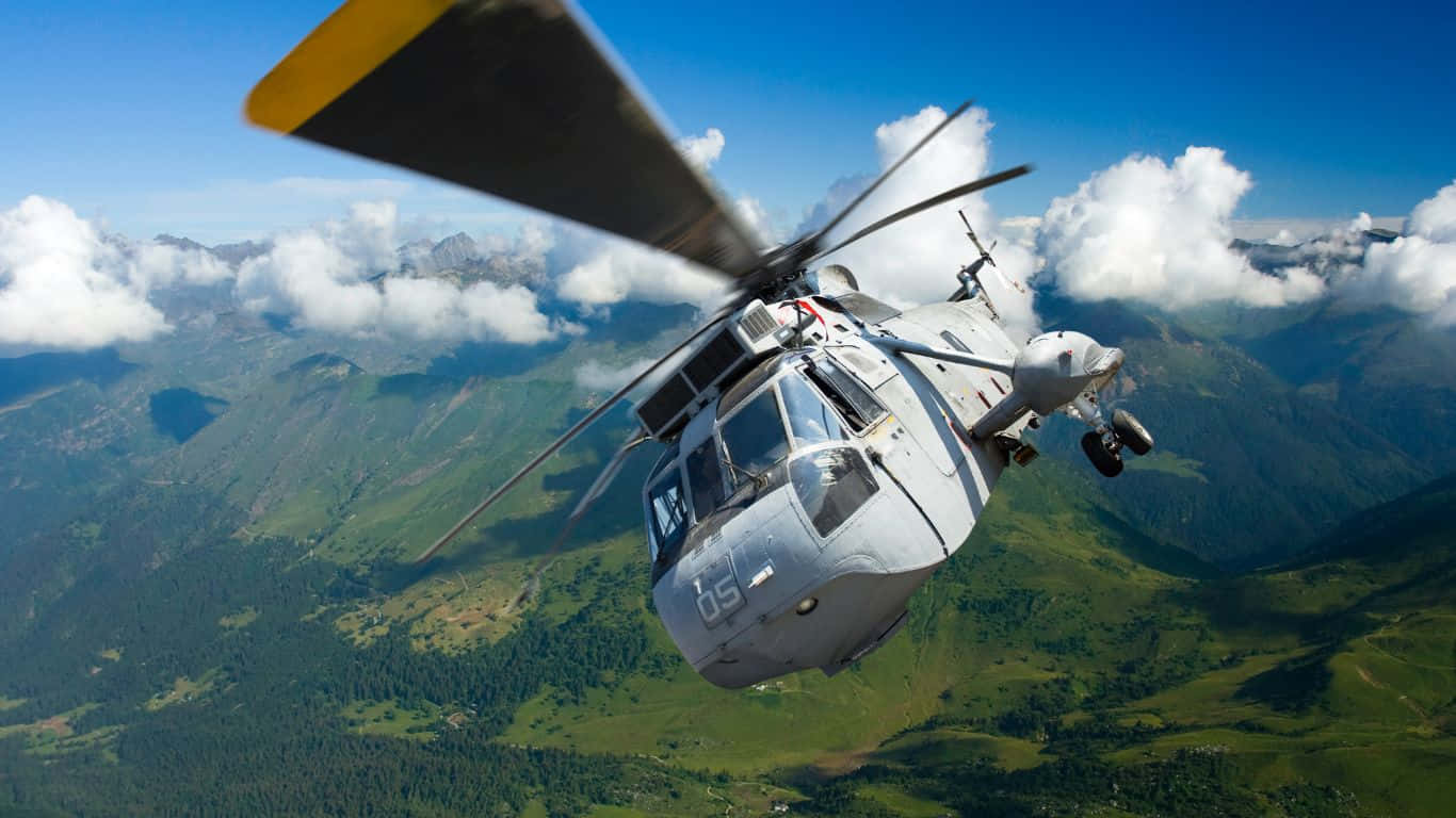 1366x768 Helicopters 桌布 壁紙 — 一架軍用直升機在點綴著蓬鬆白雲的明亮藍天下，翱翔於鬱鬱蔥蔥的綠色山脈之上。