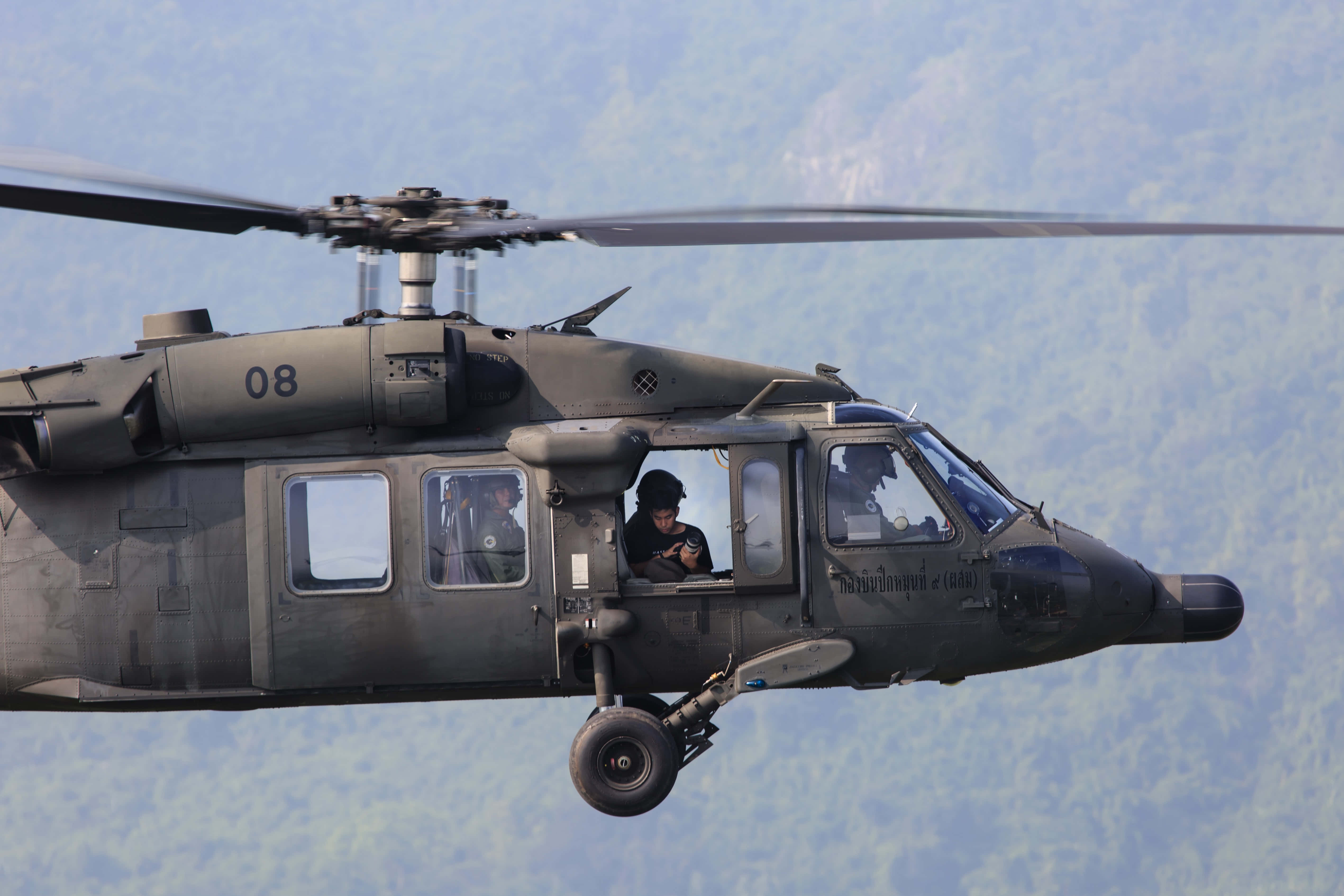 Helicopter 桌布 壁紙 — 一架軍用直升機帶有可見機組人員，在晴朗天空下低飛越過森林覆蓋的山脈。