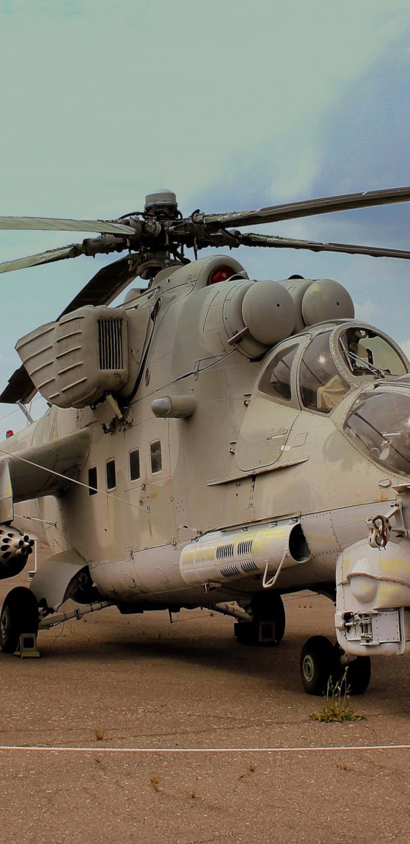 Helicopter 桌布 壁紙 — 一架軍用直升機配備旋翼和武器，停泊在塵土飛揚的停機坪上，天空多雲。