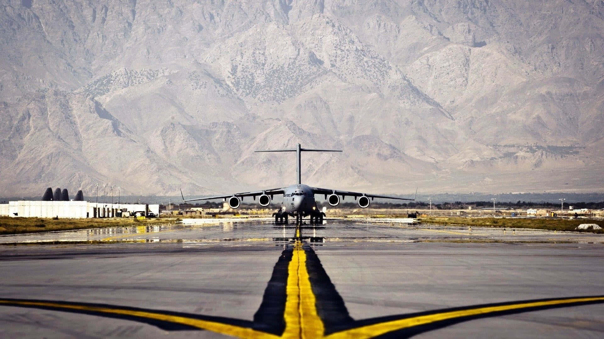 Runway 桌布 壁紙 — 一架軍用運輸機停在濕滑的跑道上，跑道有黃色中心線，遠方崎嶇的山脈在朦朧的天空下形成框架。