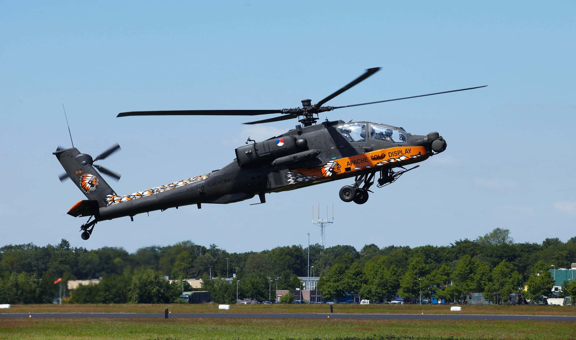 2440x1440 Helicopters 桌布 壁紙 — 一架軍用阿帕契直升機帶有橙色和黑色虎紋塗裝，在晴朗藍天下低空盤旋於跑道上空。