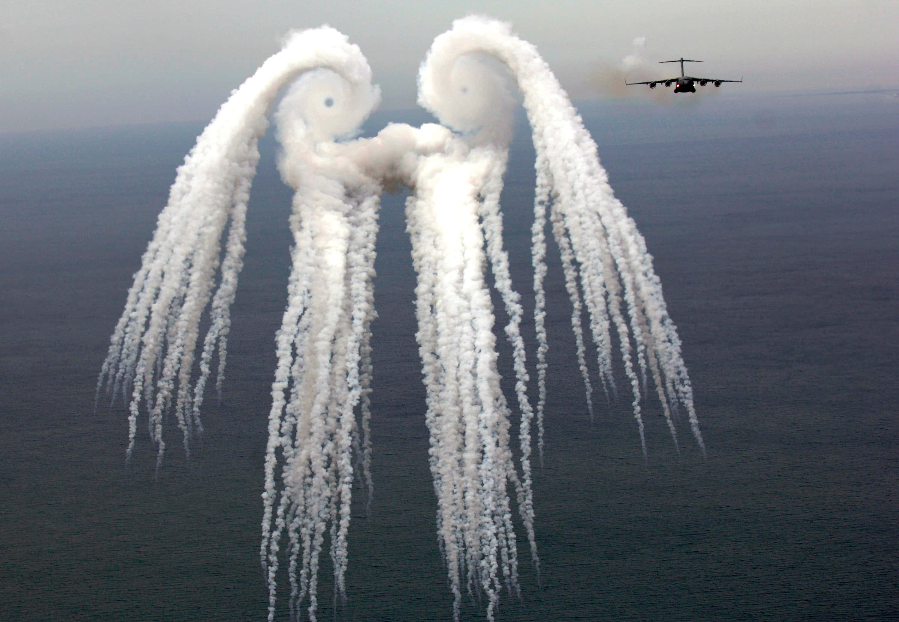 Military Plane 桌布 壁紙 — 一架軍用飛機在廣闊的海洋上空釋放出巨大的白色煙霧軌跡，形成天使翅膀。