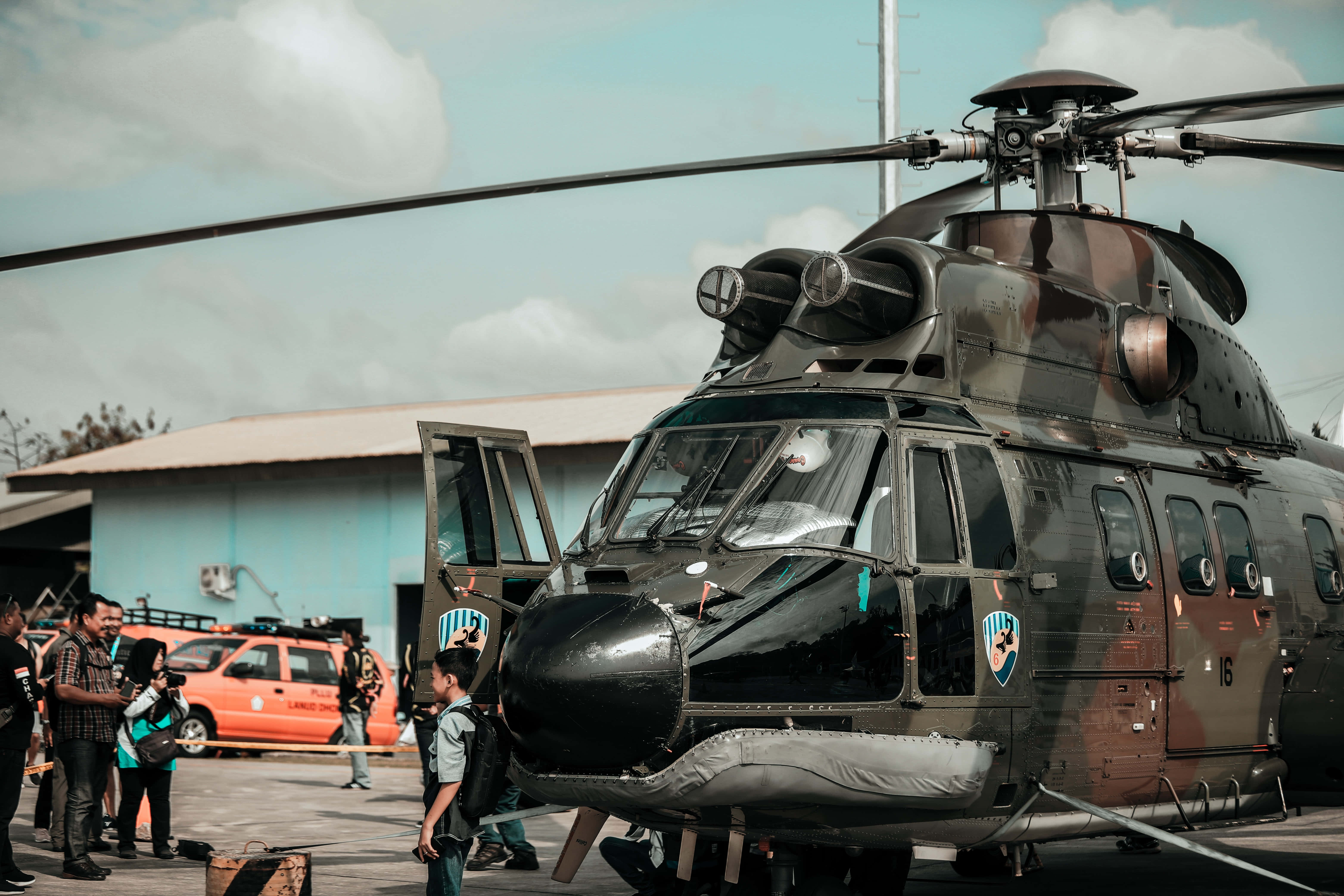 Helicopter 桌布 壁紙 — 一架迷彩塗裝的軍用直升機停在停機坪上，人們在部分多雲的天空下聚集在它周圍。
