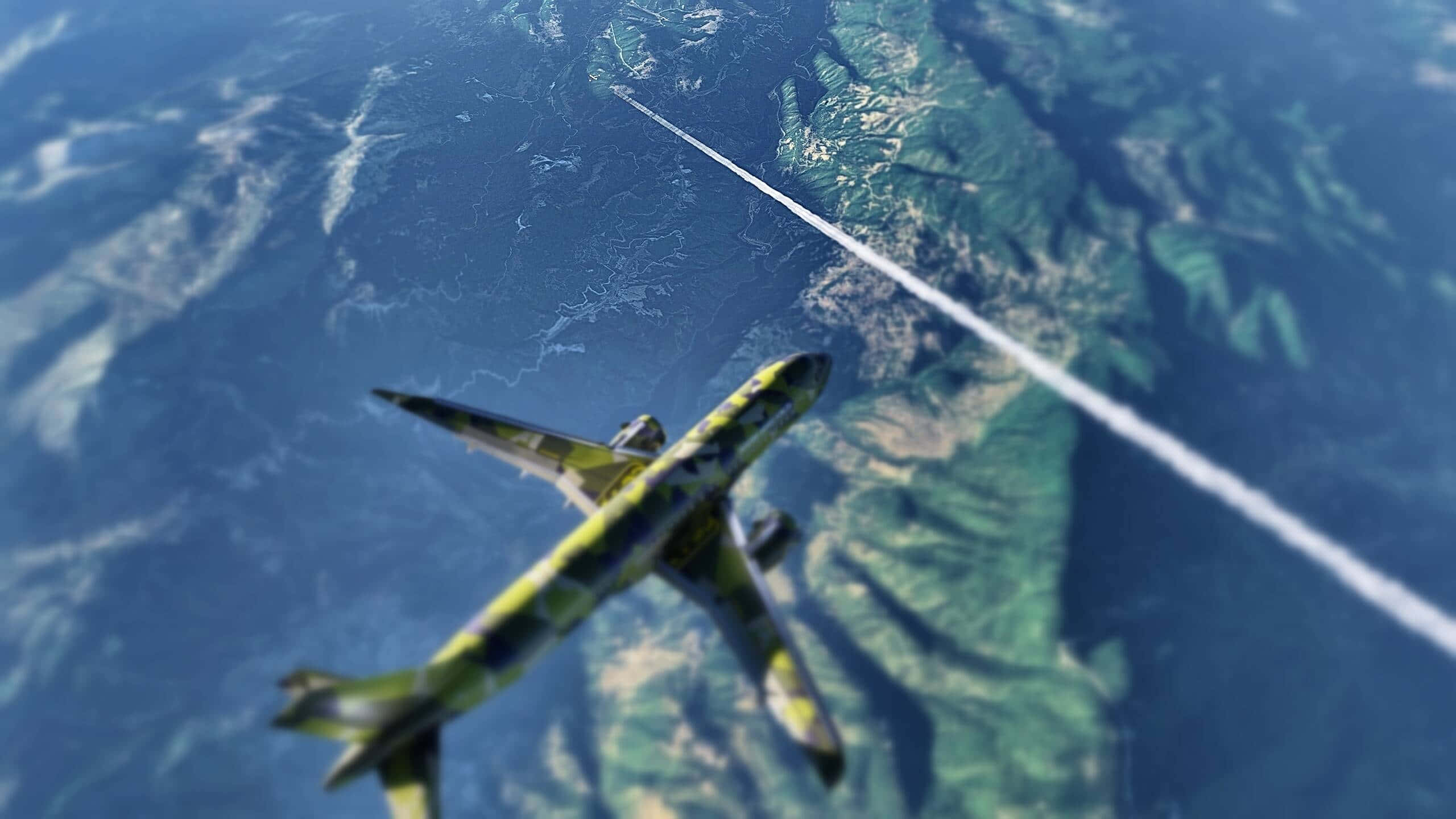 1440p Plane 桌布 壁紙 — 一架迷彩飛機翱翔於山區地形上空，在藍天中留下一道白色凝結尾跡。